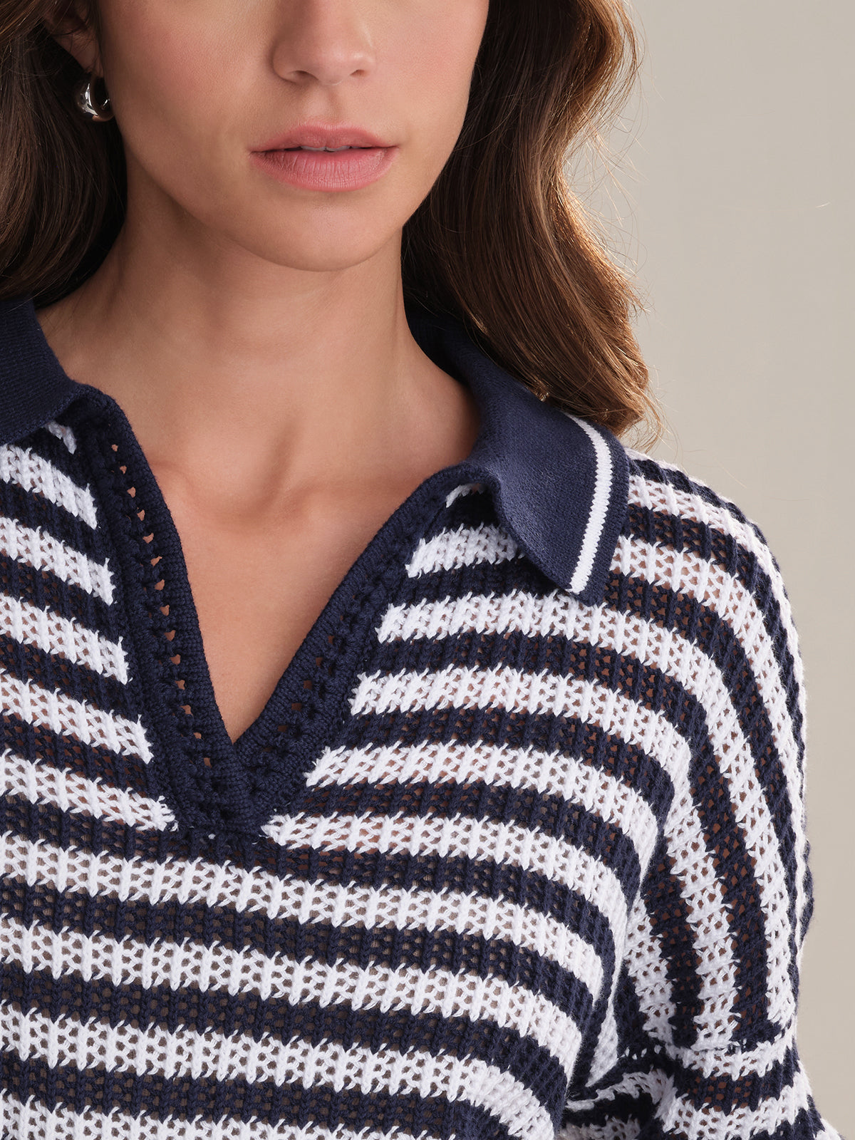 A woman models the 525 America Kimberly Stripe Polo Sweater, a navy and white cotton sweater with a polo collar and V-neckline. Only her lower face, neck, and upper torso are shown; she has long brown hair and small hoop earrings.