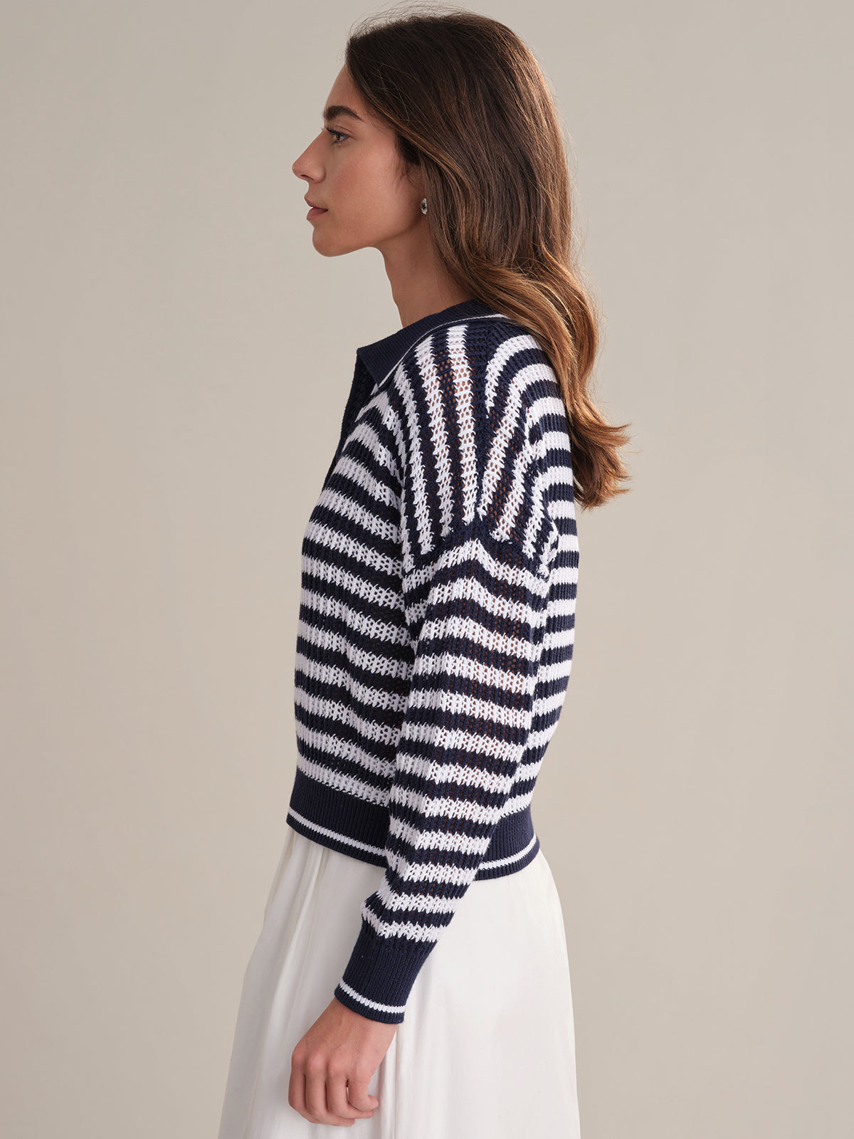 A woman with long brown hair, wearing a 525 America Kimberly Stripe Polo Sweater—navy and white cotton in a polo style—and a white skirt, stands in profile against a plain light background.