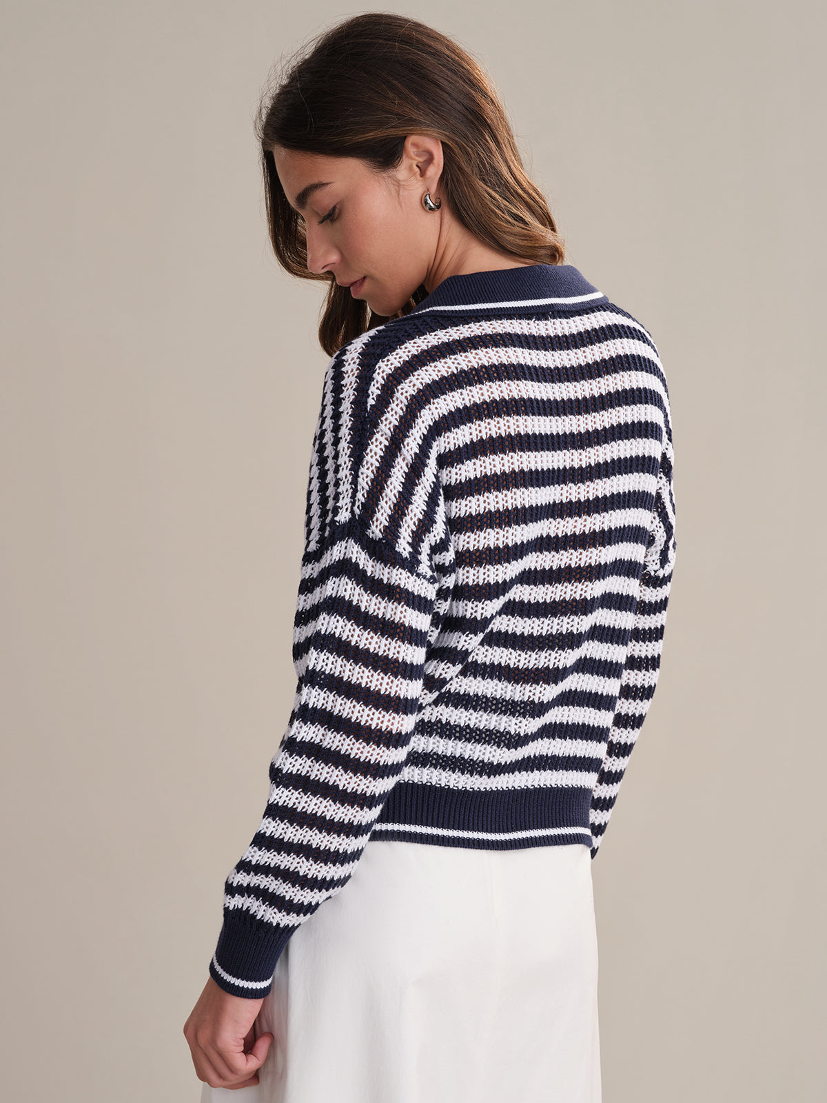 A woman with long brown hair stands facing away, wearing the 525 America Kimberly Stripe Polo Sweater—navy and white cotton in a classic style—paired with a white skirt, against a plain neutral background.