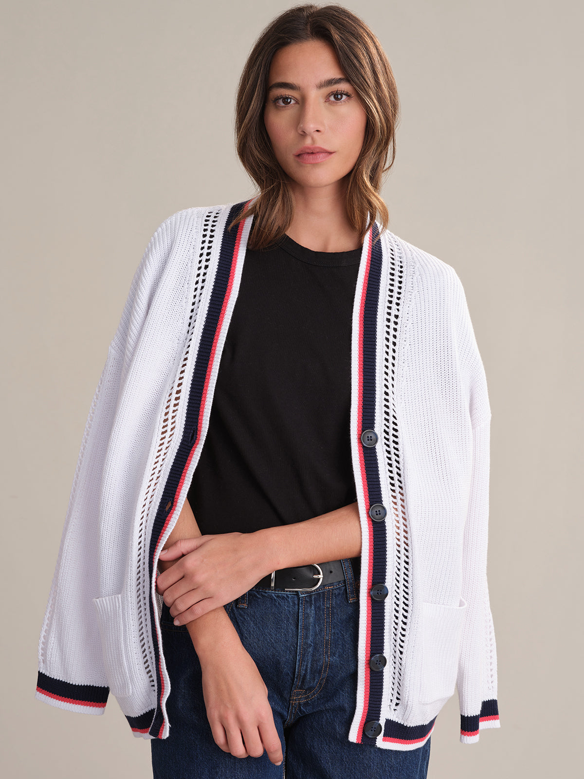 A woman with wavy brown hair and a neutral expression wears a 525 America relaxed-fit cotton cardigan featuring red, navy, and white varsity-style striped trim over a black t-shirt and blue jeans, standing against a plain background.