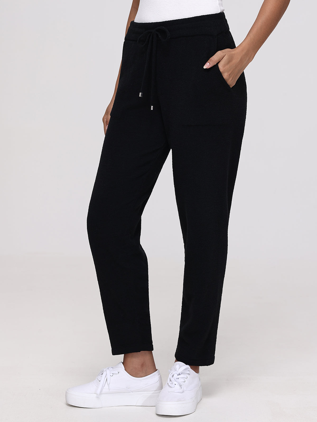 A person in black drawstring jogger pants by 525 America, made from plush fabric, and white sneakers stands against a plain background. They complete the look with one hand in a pocket and a white top.