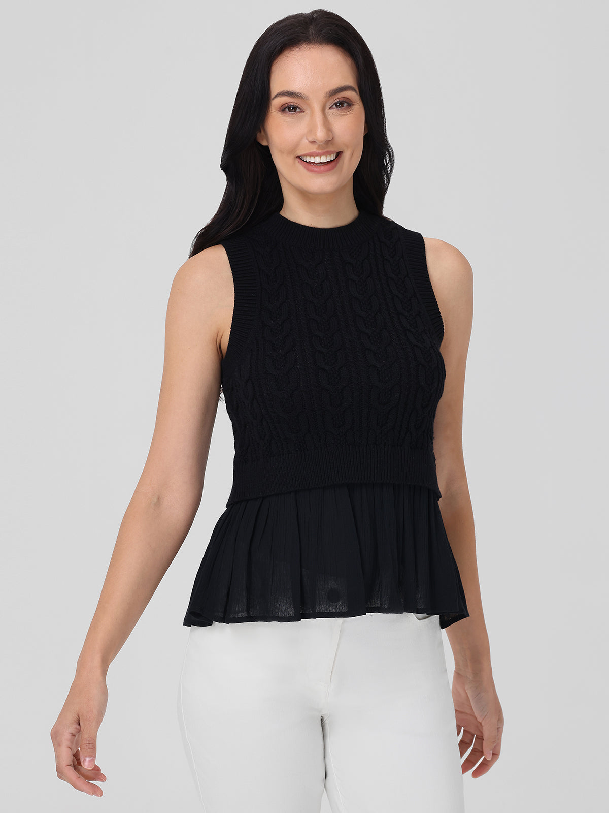 A person with long dark hair wears a 525 America black sleeveless textured knit top, ideal for layering, featuring a sheer pleated lower section. Its paired with white pants and stands out against a plain light gray background.