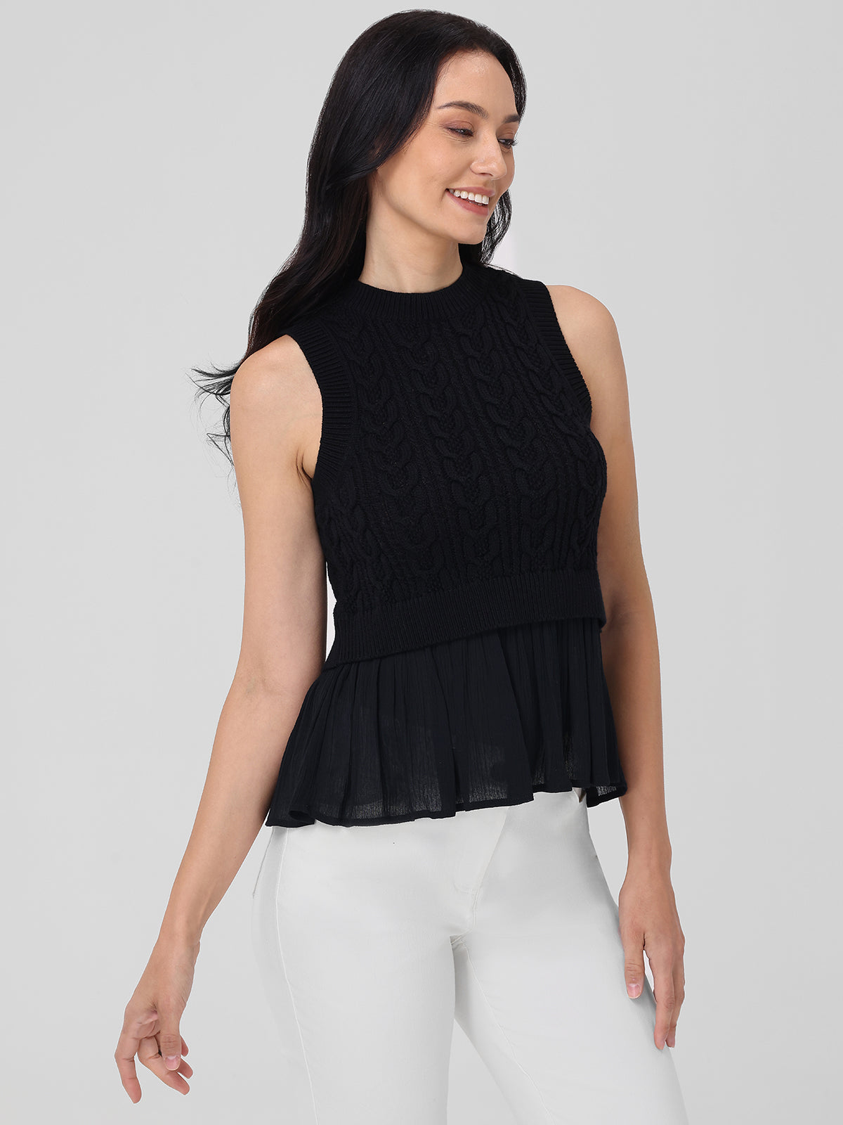 A person with long dark hair wears a 525 America black sleeveless knit top and white pants, a versatile seasonal staple. The outfits pleated peplum hem is great for layering. They stand against a plain gray background, smiling and looking to the side.