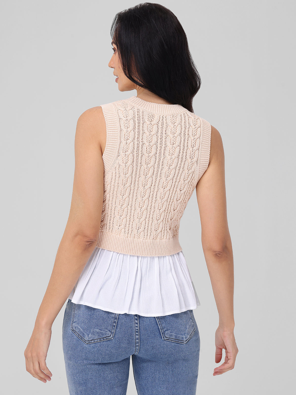 A person with long dark hair is seen wearing a sleeveless beige knitted top by 525 America, made of 100% cotton and featuring white pleated fabric at the bottom—a seasonal staple with textured patterns. They pair it perfectly with blue jeans against a plain gray background, facing away.