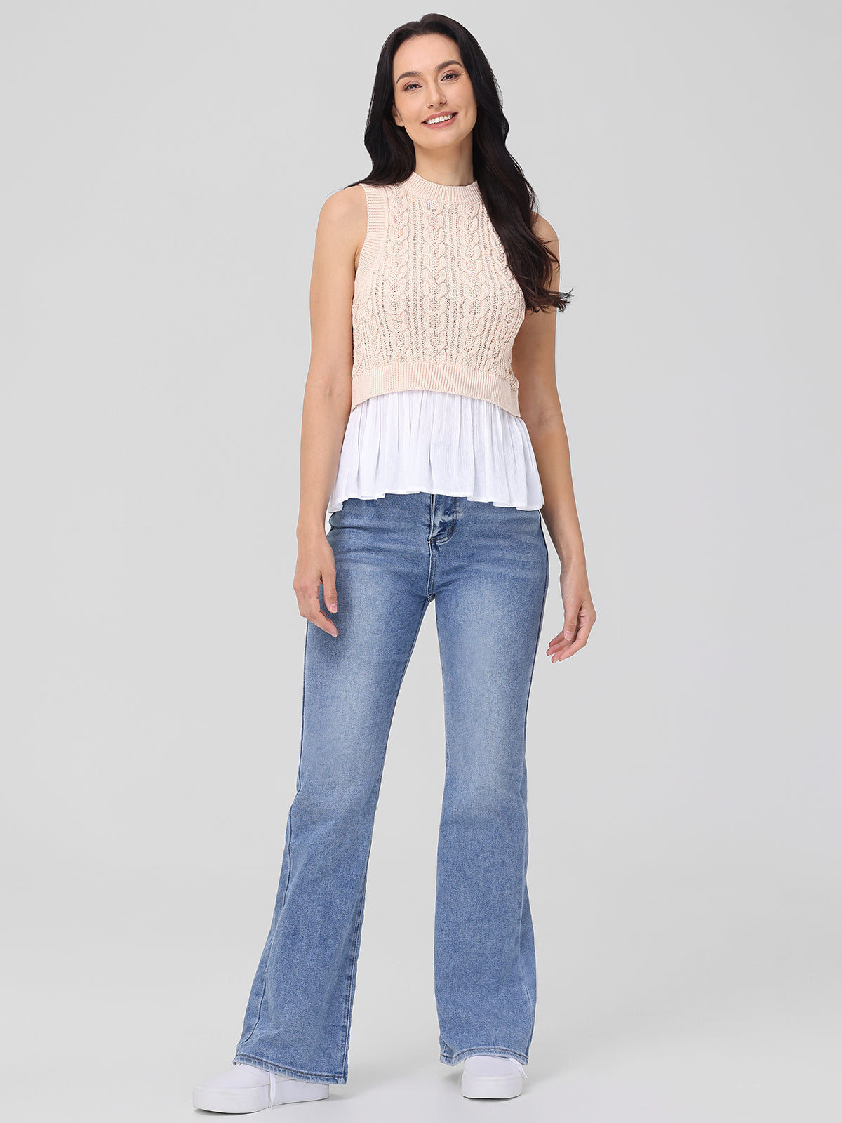A person with long dark hair poses against a simple backdrop, showcasing seasonal staples by 525 America. They wear a sleeveless beige knit top over a white flowy blouse, paired with light blue flared jeans and white sneakers. Smiling, they keep their arms relaxed by their sides.