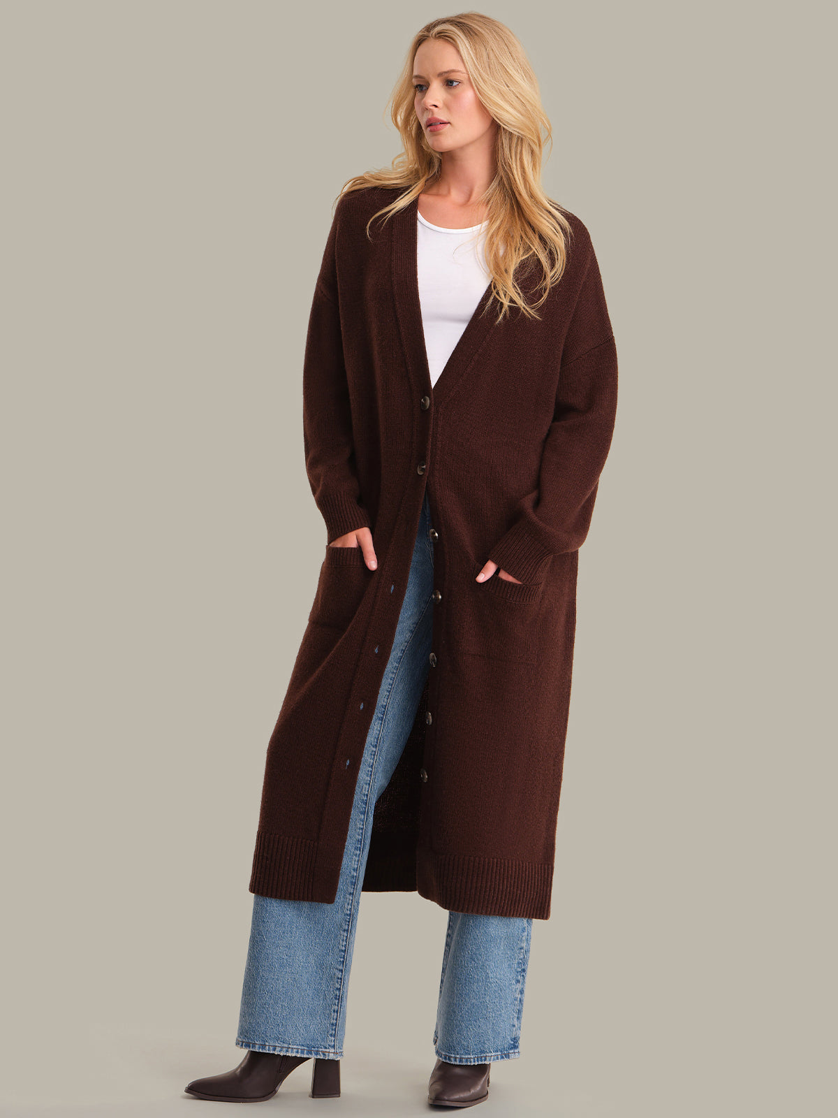 Woman wearing a long brown coat over a white top and blue jeans on a gray background