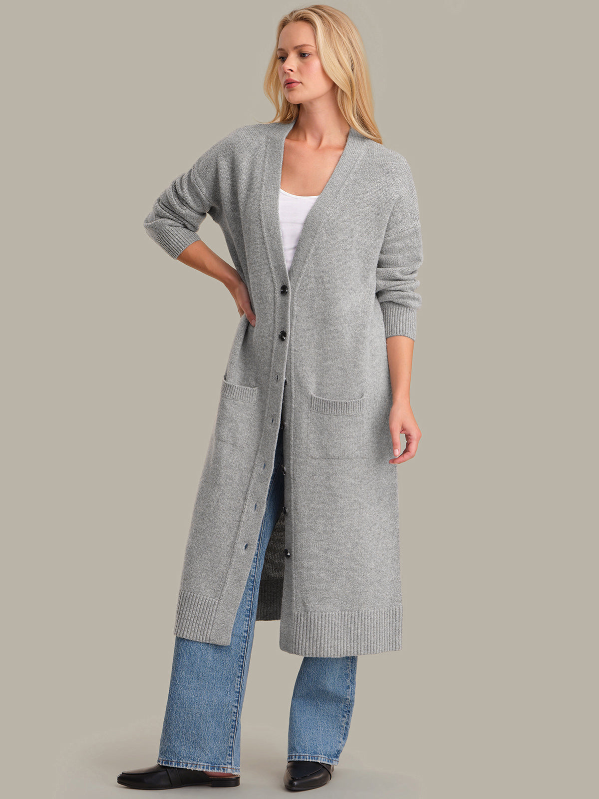 Woman wearing a long gray cardigan over a white top and blue jeans on a plain background