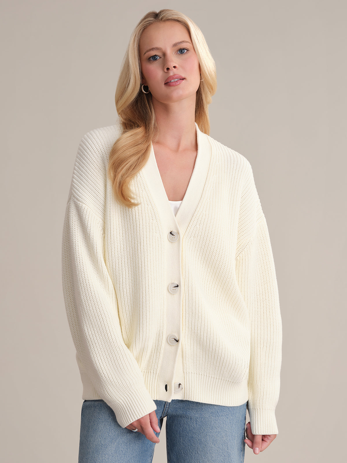 A person with long blonde hair is wearing the Emilia: Cotton Cocoon Cardigan, a cream-colored, oversized 100% cotton design with large buttons. Its layered over a white top and blue jeans against a plain background.