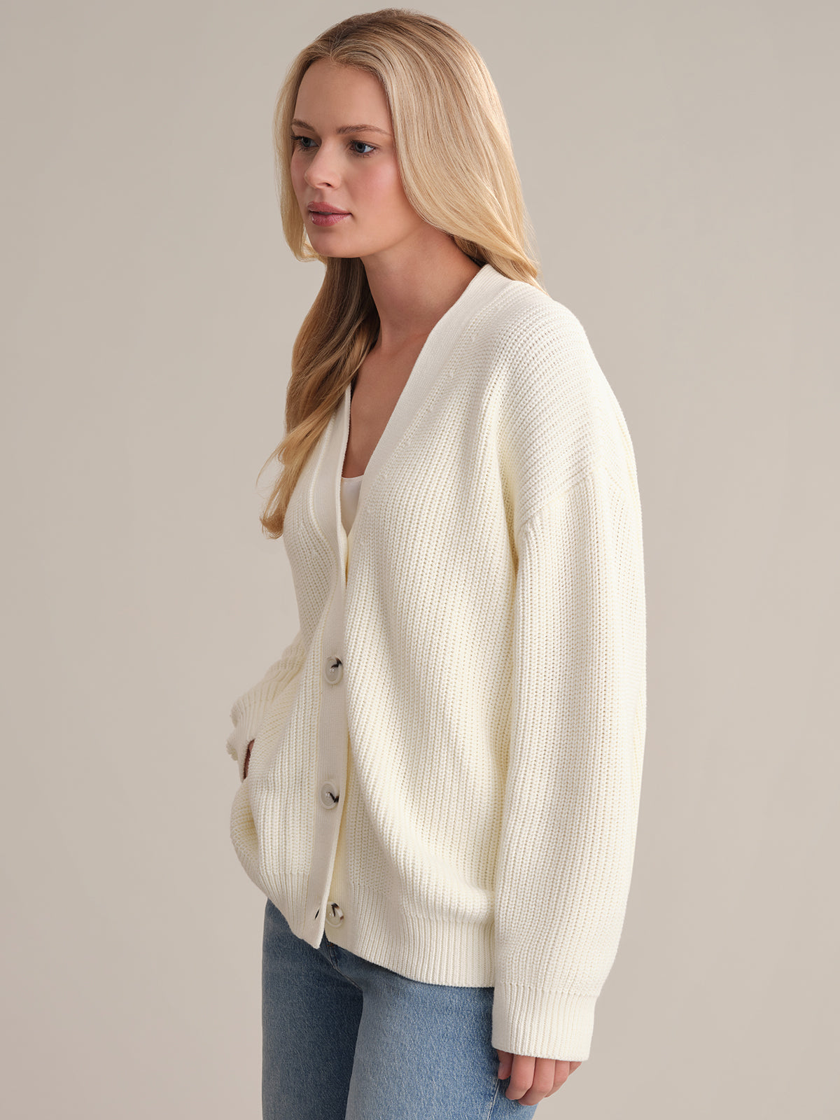 A person with long hair wears an oversized Emilia: Cotton Cocoon Cardigan in white over a light shirt and blue jeans. They stand against a plain backdrop, looking to the side, effortlessly mastering the art of layering.