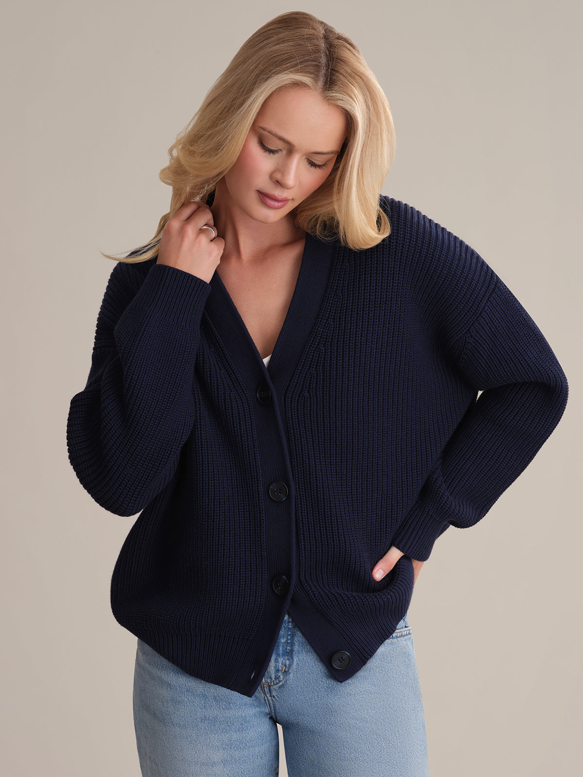 Someone with long blonde hair wears the Emilia: Cotton Cocoon Cardigan in dark blue, in an oversized fit, paired with light blue jeans. They look down, one hand on their neck and the other on their hip against a neutral background, showcasing a cozy yet stylish layered look.