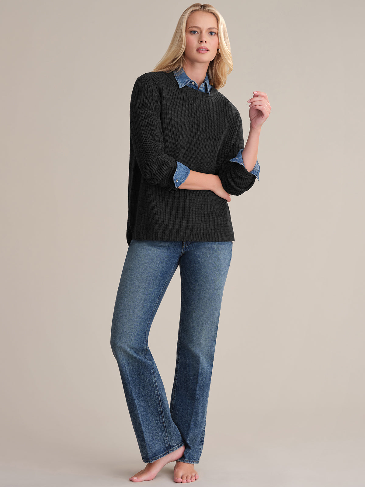A person with long hair wears the Emma: Wool Crewneck Shaker Stitch Sweater in black, layered over a denim shirt and paired with blue jeans. They stand barefoot against a plain background, posing casually with one hand slightly raised.
