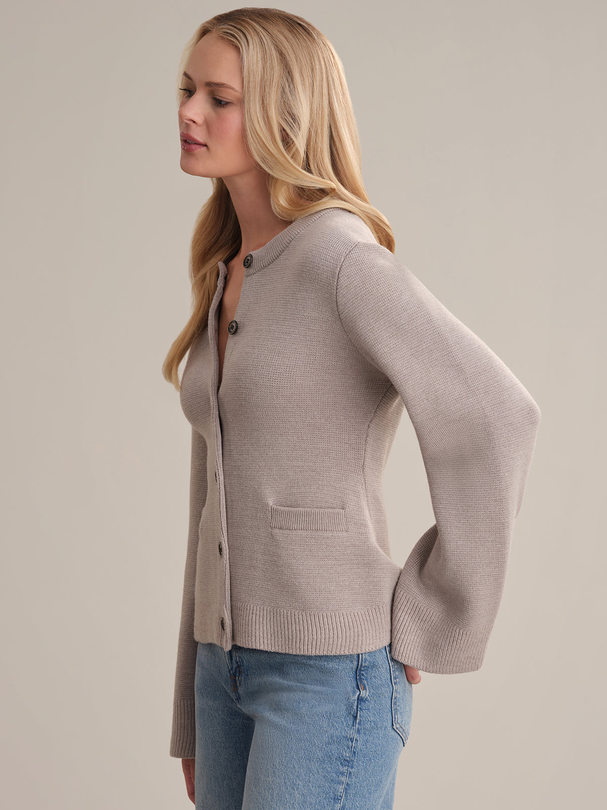 A person with long blonde hair is seen in profile, wearing the elegant Catherine wool cardigan with pockets and blue jeans against a plain neutral background.