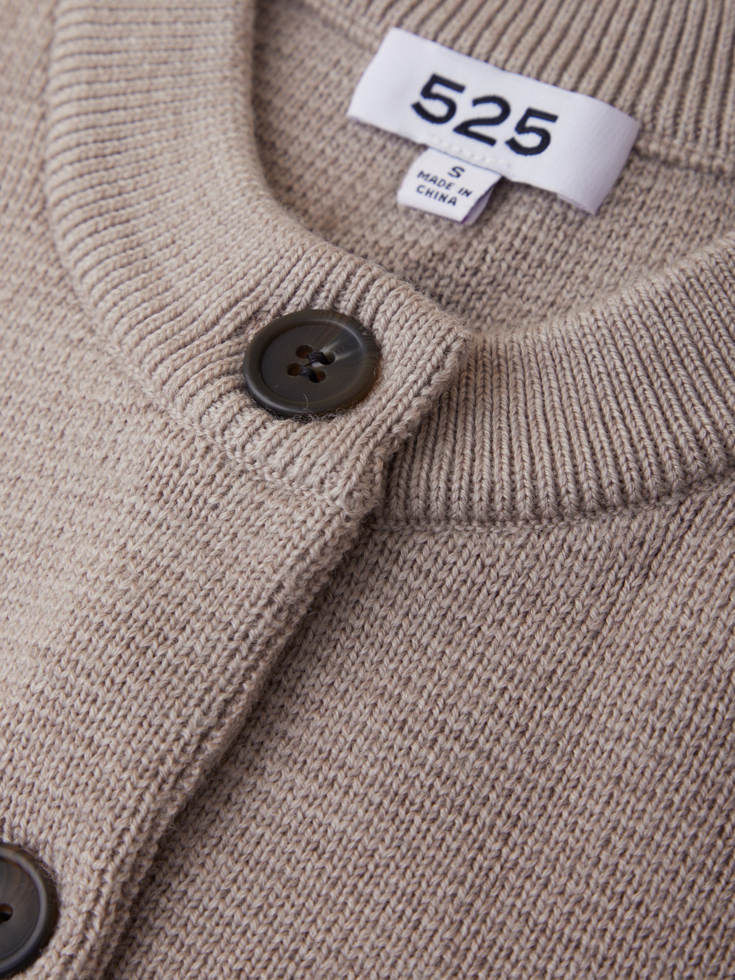 Close-up of a beige cardigan with a button and brand label on a plain background