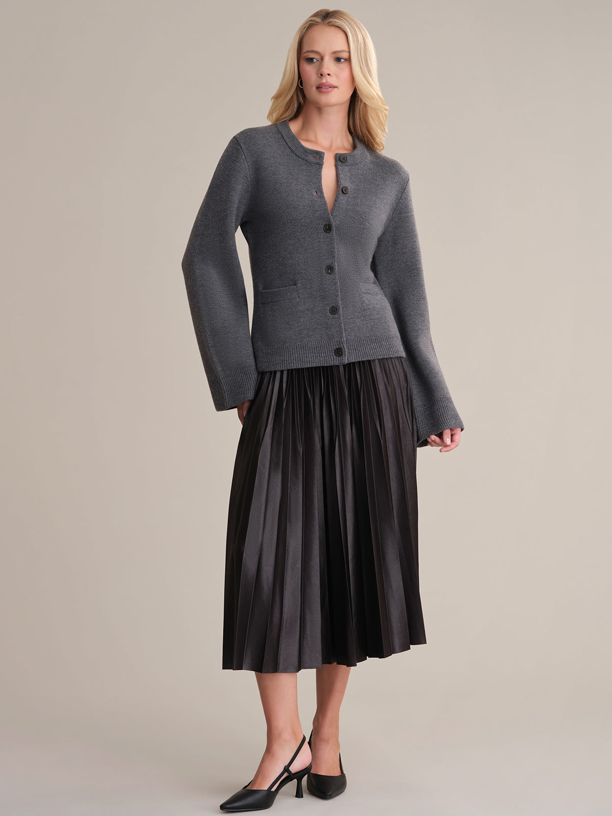 A woman with long blonde hair exudes sophistication in the Catherine gray wool cardigan and a black pleated midi skirt, pairing her look with black heeled shoes. The plain background highlights her chic style.