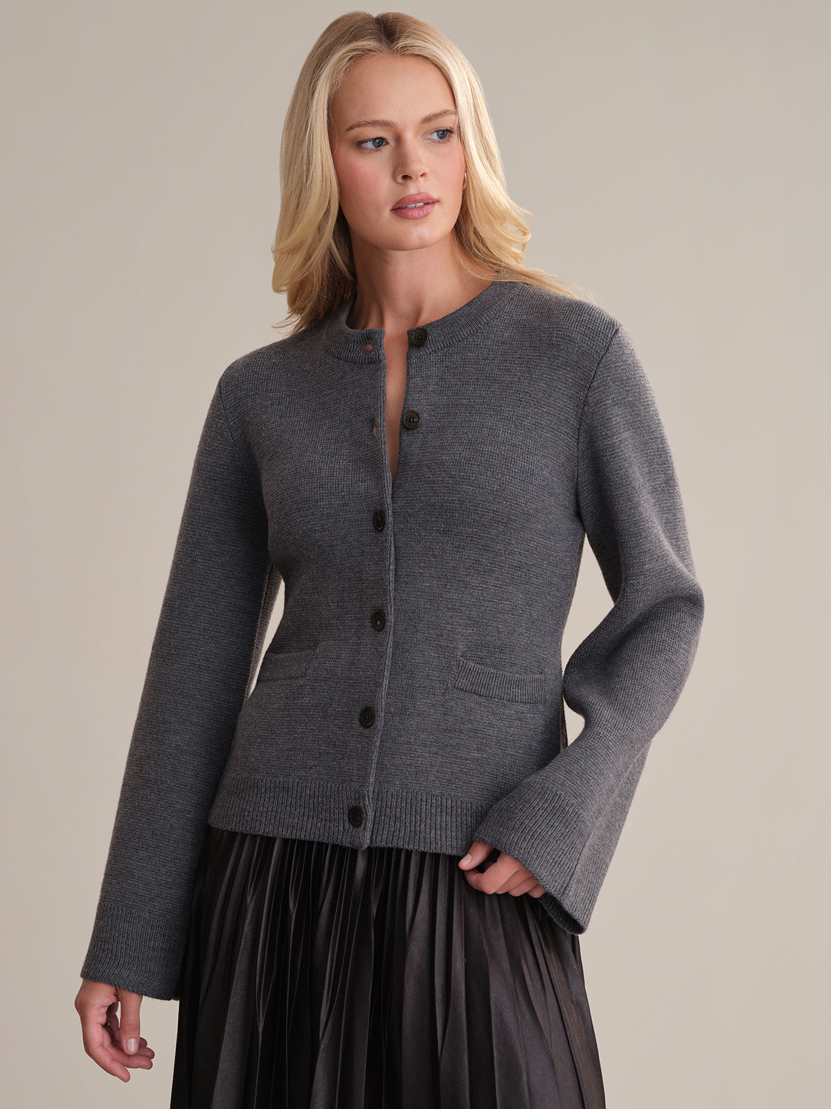 Graced by a backdrop, a woman emanates elegance in the Catherine wool cardigan with black buttons. Her long blonde hair cascades as she gazes aside, her look flawlessly completed by a dark pleated skirt.