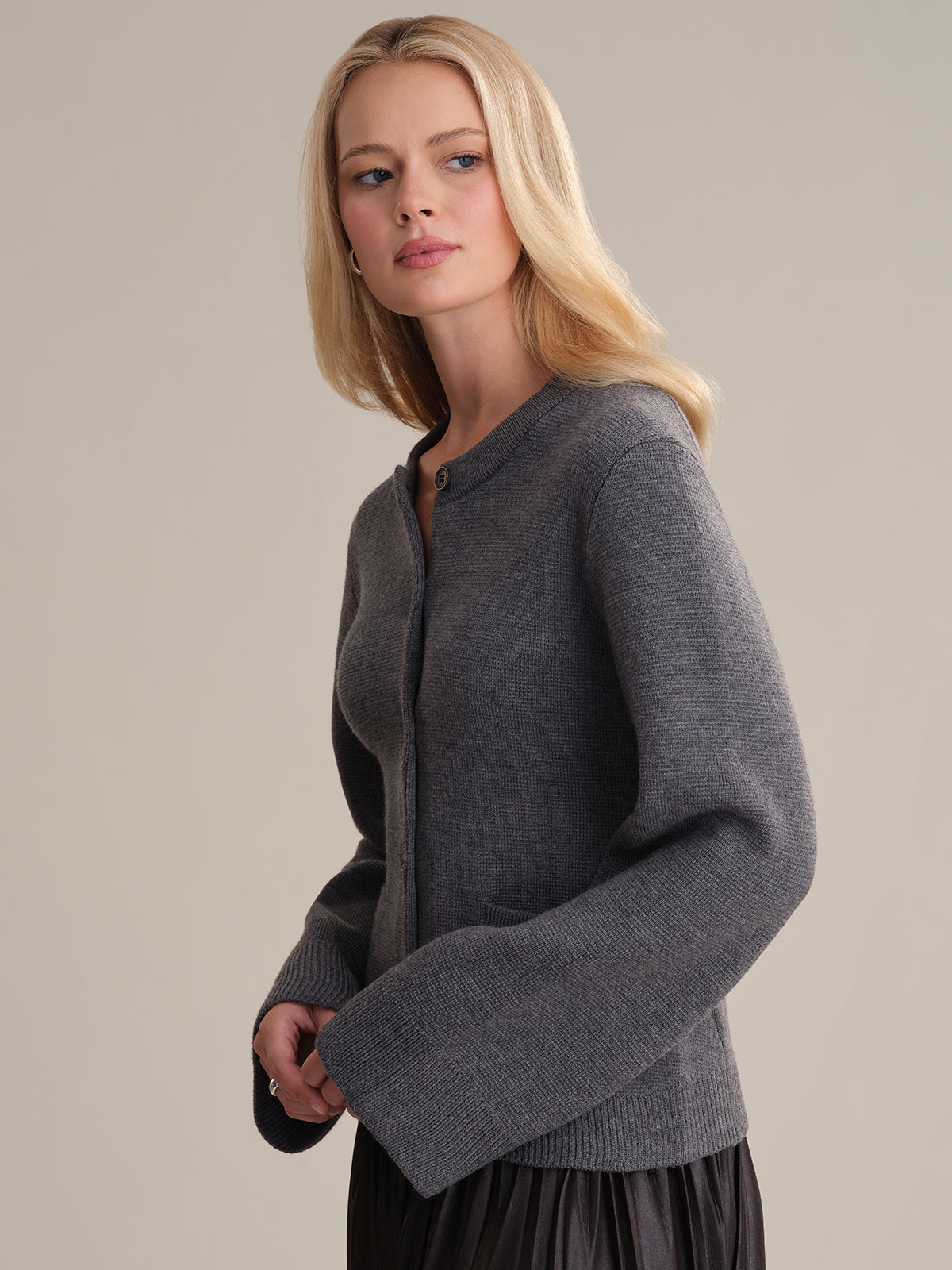 A person with long blonde hair exudes sophistication in the Catherine: Wool Cardigan featuring wide sleeves and a chic black pleated skirt, set against a simple backdrop. They gaze slightly to the side with a neutral expression.