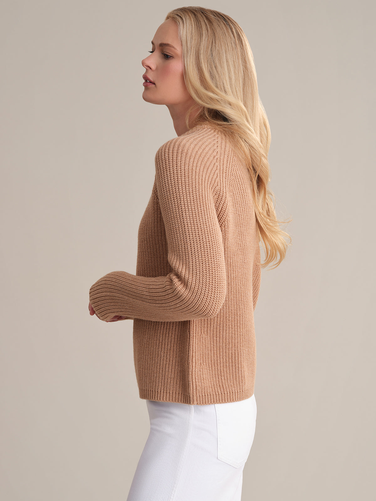 A person with long blond hair stands sideways in a Jane Crewneck Shaker Stitch Cotton Sweater with sporty raglan sleeves, paired with white pants, against a neutral background.