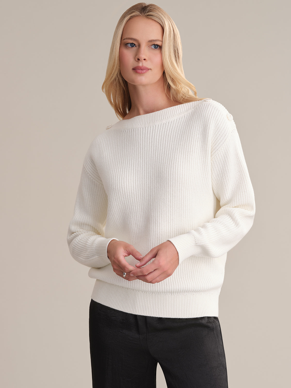 A person with long blonde hair wears a versatile white ribbed sweater by 525 America featuring a boat neck, paired with black pants. They stand against a plain background, clasping their hands at waist level and looking slightly to the side.