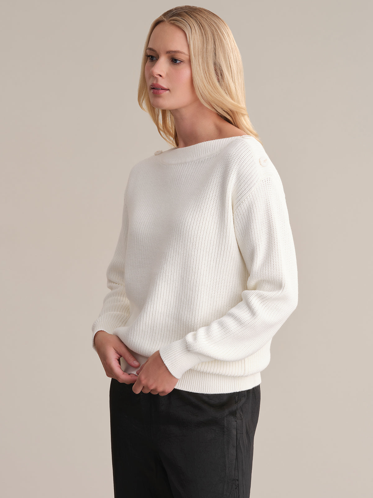A person with long blonde hair stands against a plain beige background, wearing a versatile white ribbed boat neck sweater by 525 America and black pants, looking to the side with a neutral expression.