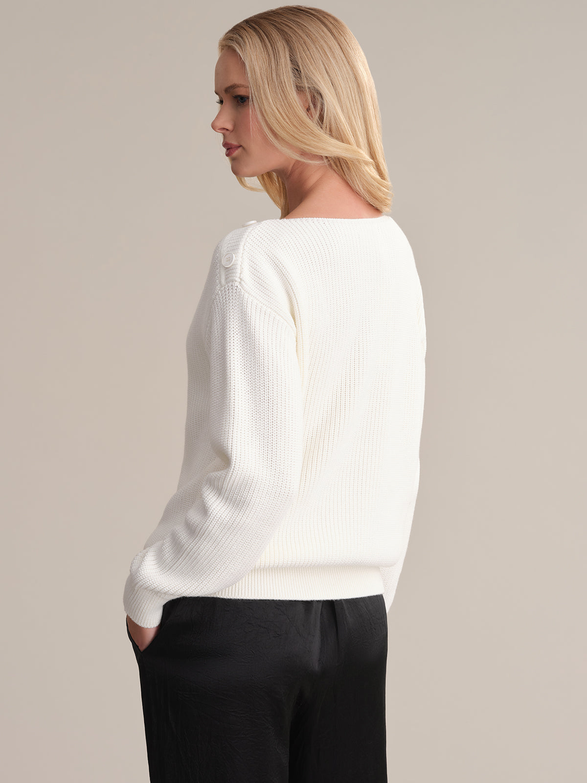 A person with long blonde hair stands sideways, looking ahead in a versatile 525 America sweater with a boat neck. They effortlessly pair it with black pants, hands in pockets, against a neutral backdrop that complements the chic outfit.