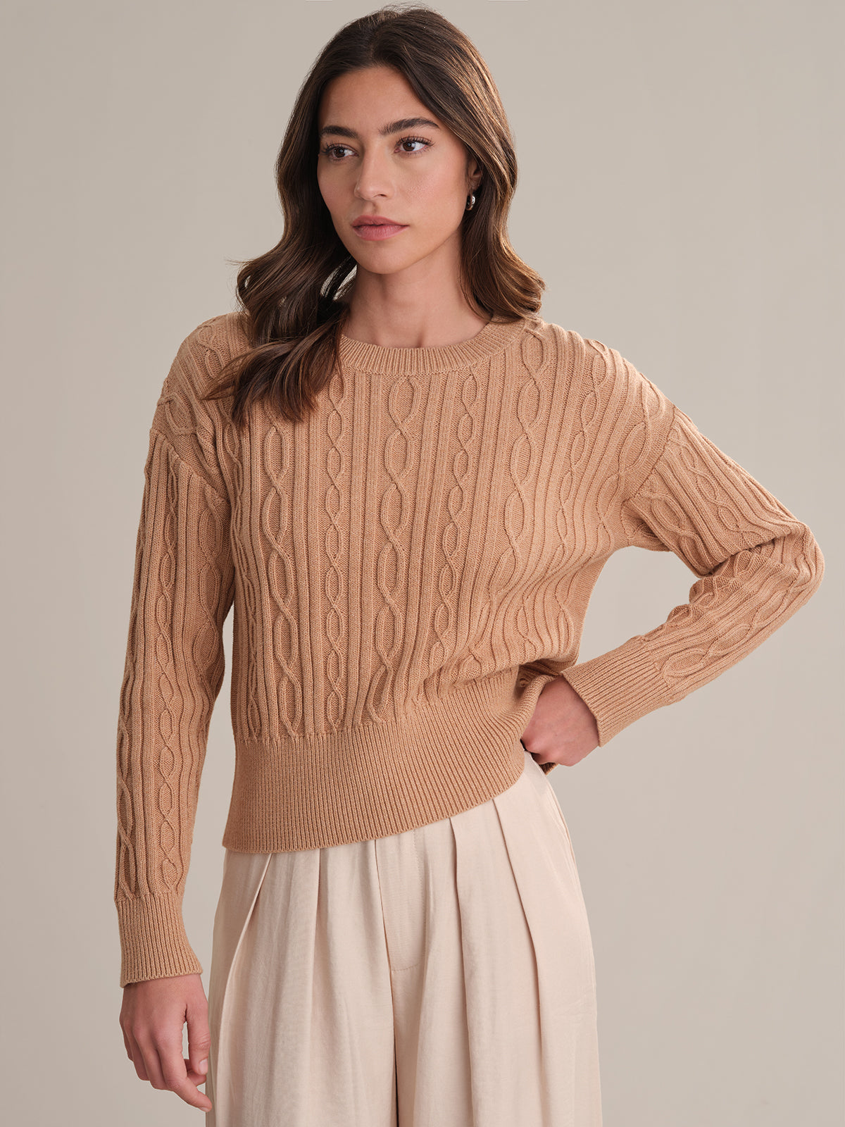 A woman with long brown hair models a tan 525 America Cable Cotton Sweater with classic cable knit, paired with light beige high-waisted pants. She stands before a neutral backdrop, hand on her hip and gazing slightly to the side.