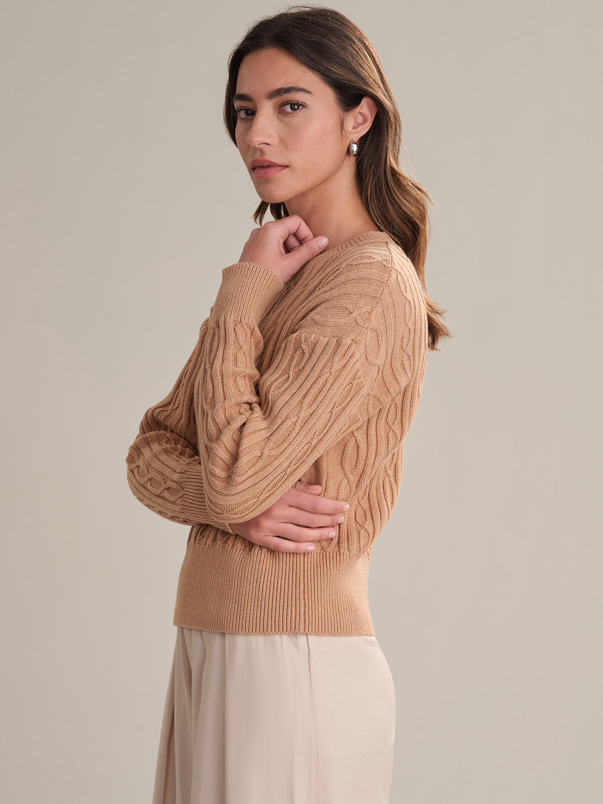 A woman with long brown hair stands in profile against a plain background, wearing a 525 America cable cotton sweater with a crewneck and a beige skirt. She gazes slightly toward the camera with a neutral expression.