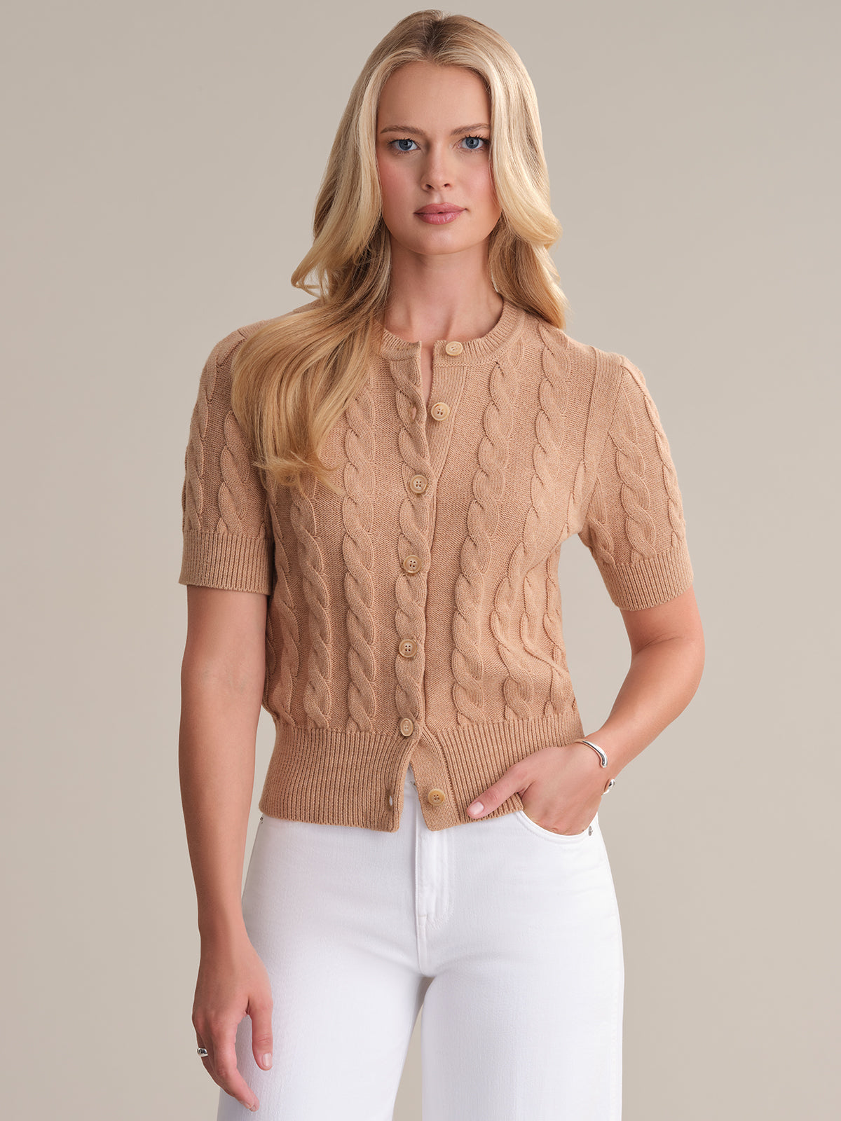 A person with long, wavy hair is elegantly wearing a short-sleeved, beige cable-knit cardigan from 525 Americas latest seasonal colorways collection. They pair it with white pants and stand gracefully against a plain, light-colored background.