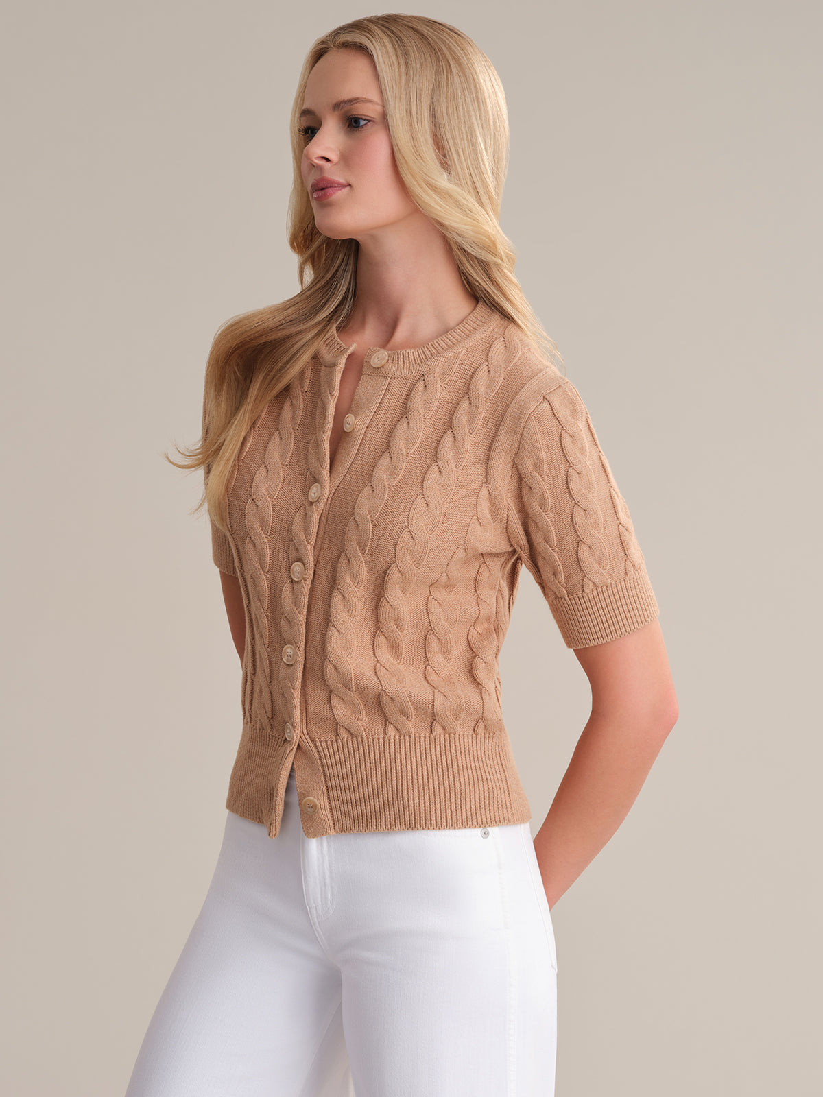 A person with long blonde hair is wearing a beige cable-knit short-sleeve sweater from 525 America and white pants. They stand against a neutral background, showcasing one of the seasonal fashion colorways while facing slightly to the side.