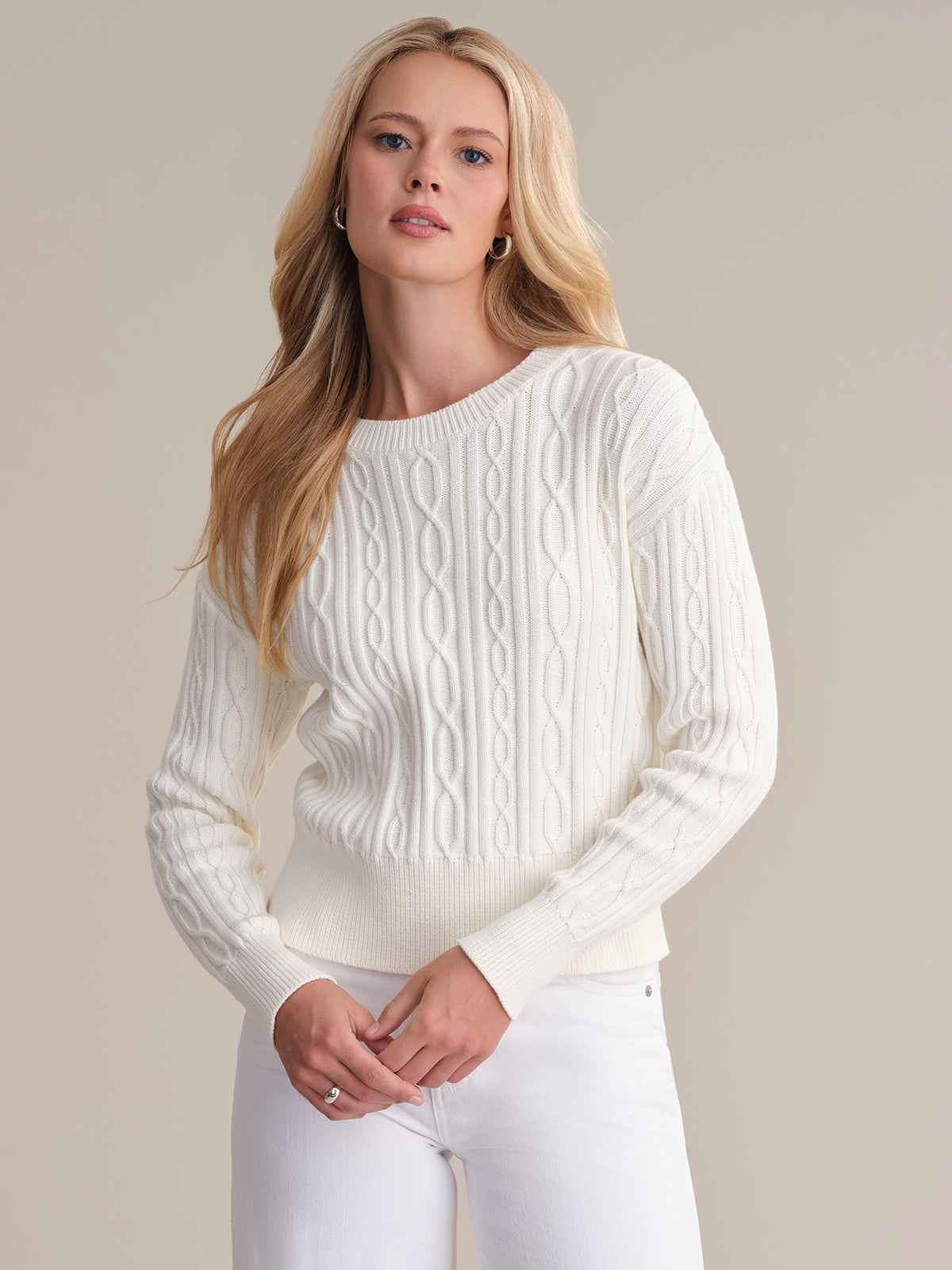 A person with long blonde hair wears a 525 America classic cable-knit sweater made of 100% cotton paired with white pants, standing against a plain, neutral background and looking slightly to the side.