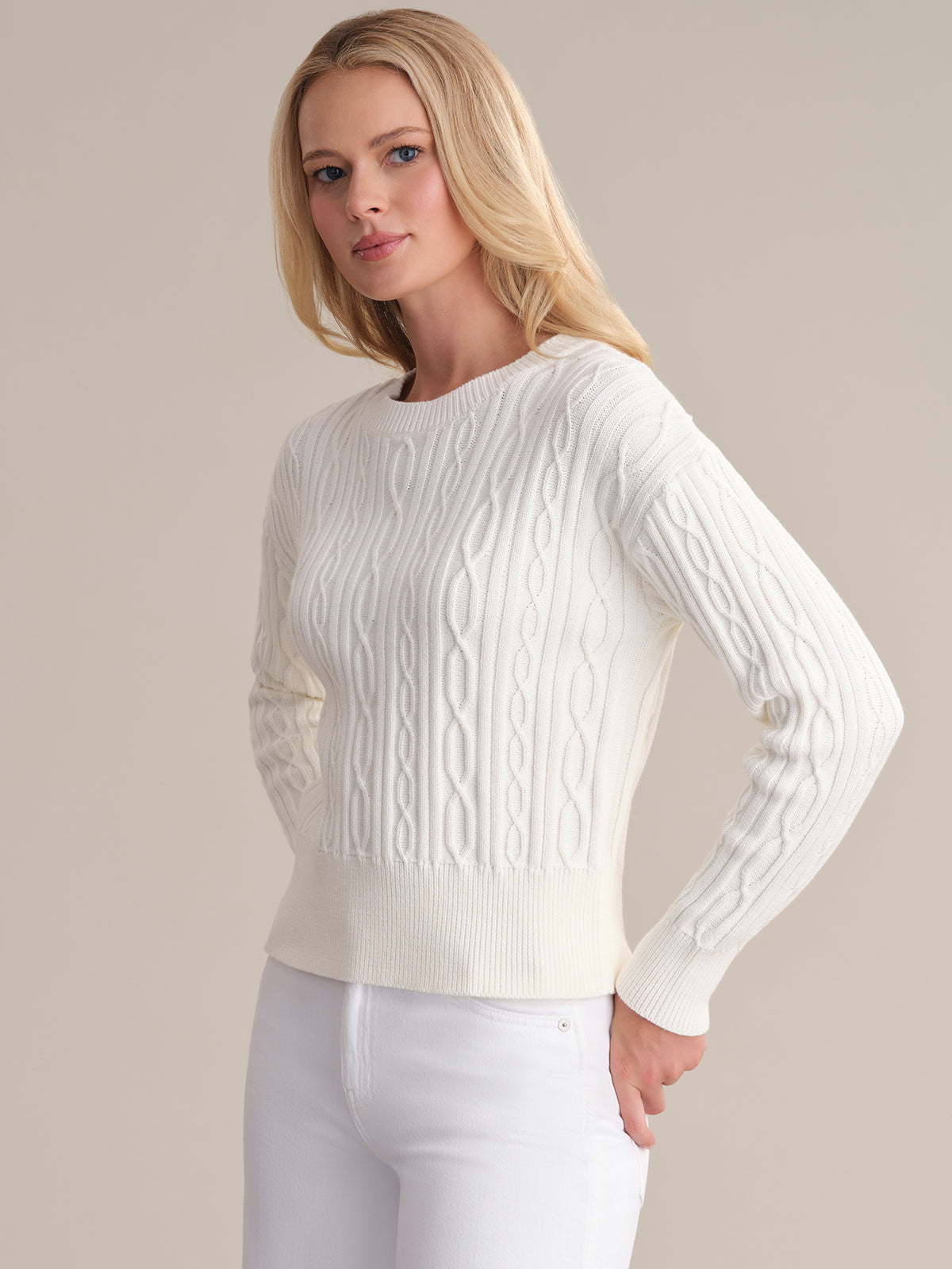 A person with long blonde hair wears a classic cable cotton sweater from 525 America and white pants against a neutral background. The 100% cotton fabric heightens the timeless appeal of their look.