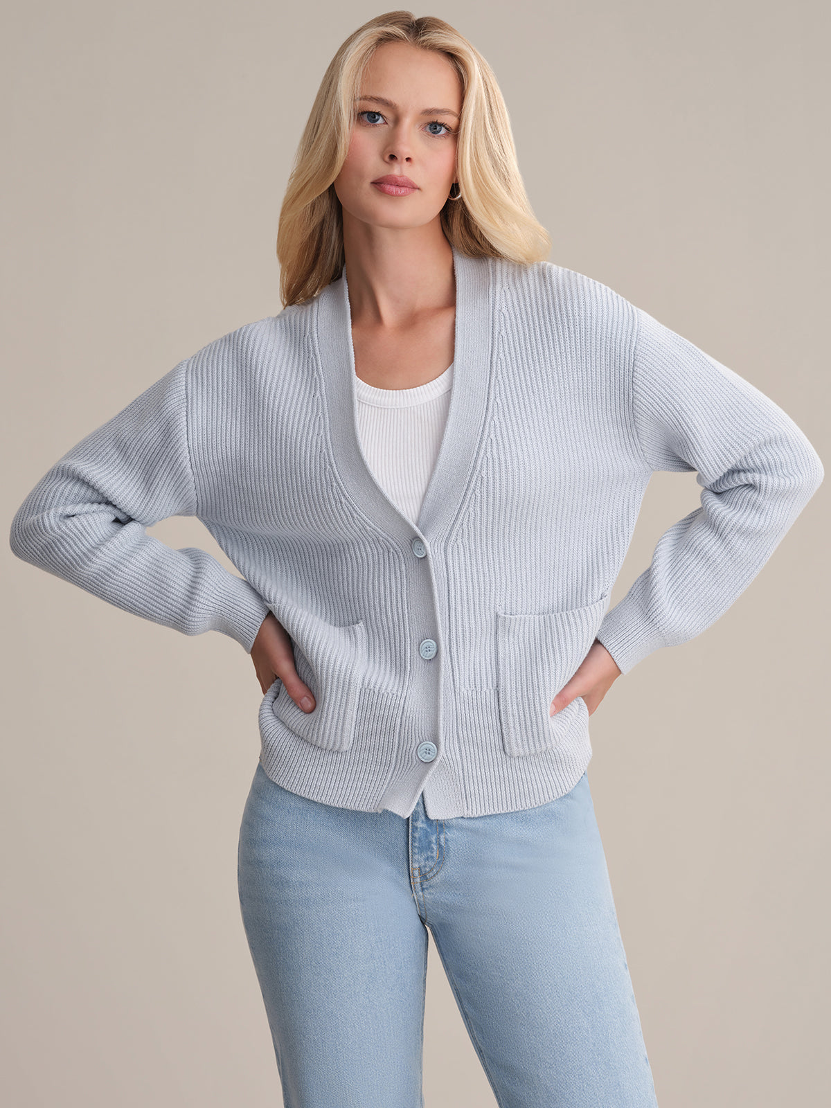 A woman with long blonde hair wears a light blue 525 America cotton cardigan over a white top and light blue jeans, standing with her hands on her hips against a plain background.