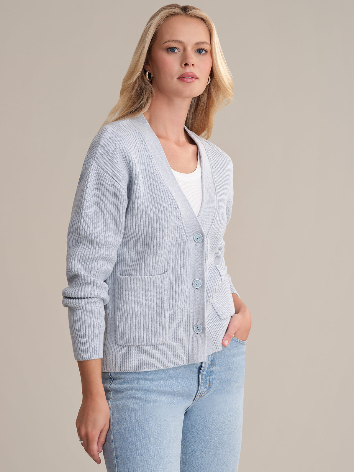 A woman with long blonde hair models a 525 America light blue cotton cardigan over a white top and matching jeans, standing with one hand in her pocket against a plain beige background.