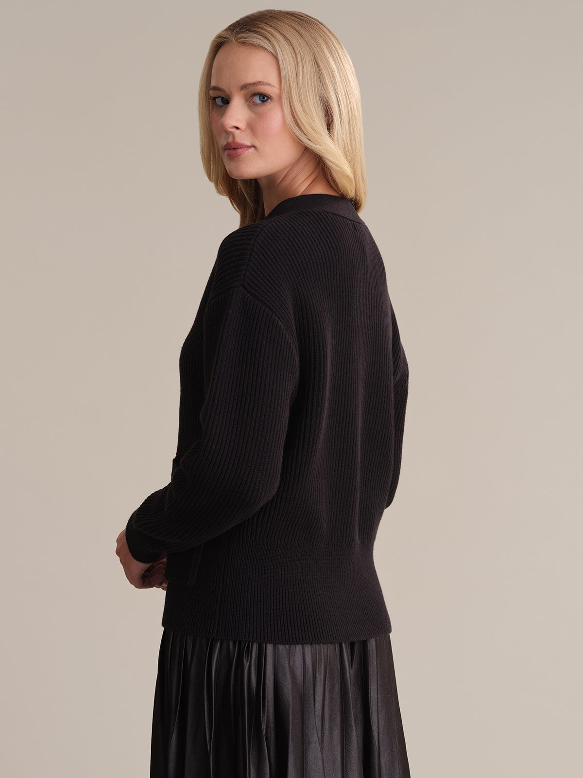 A person with long blond hair wears a 525 America black ribbed sweater, a dark pleated skirt, and a stylish v-neck cardigan. They stand sideways, glancing over their shoulder against a plain, neutral background.