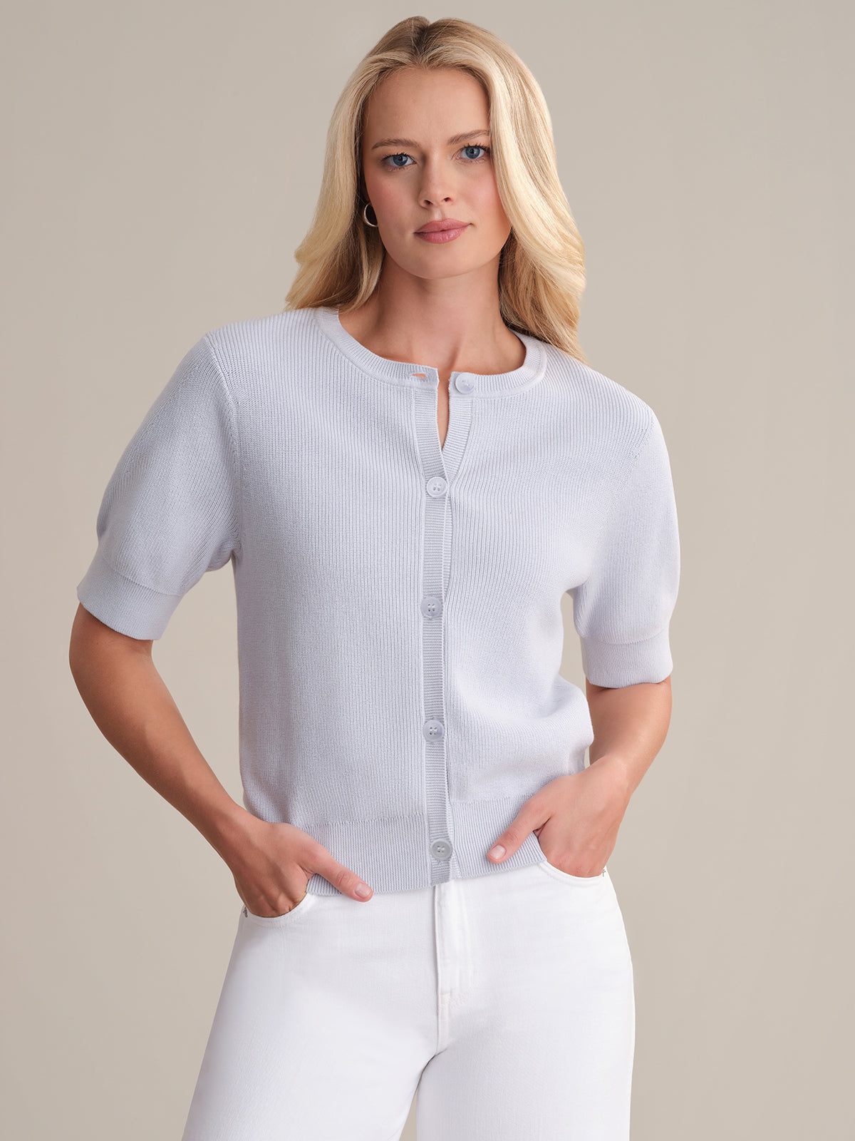 A woman with long blonde hair wears a 525 America Short Sleeve Puff Cardigan in soft cotton with large buttons and white pants, posing against a beige background, hands in pockets.
