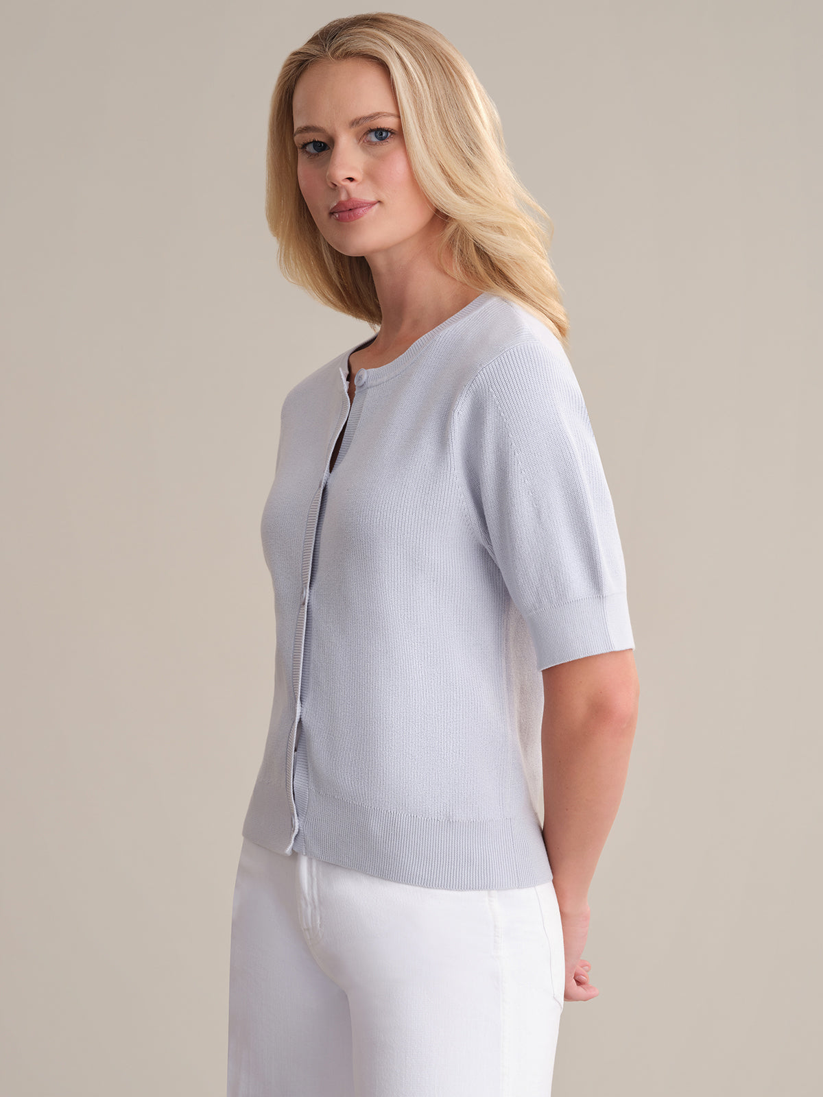 A blonde woman wears a light gray 525 America Short Sleeve Puff Cardigan made from soft cotton, paired with white pants. She stands against a plain light background, gazing slightly to the side.