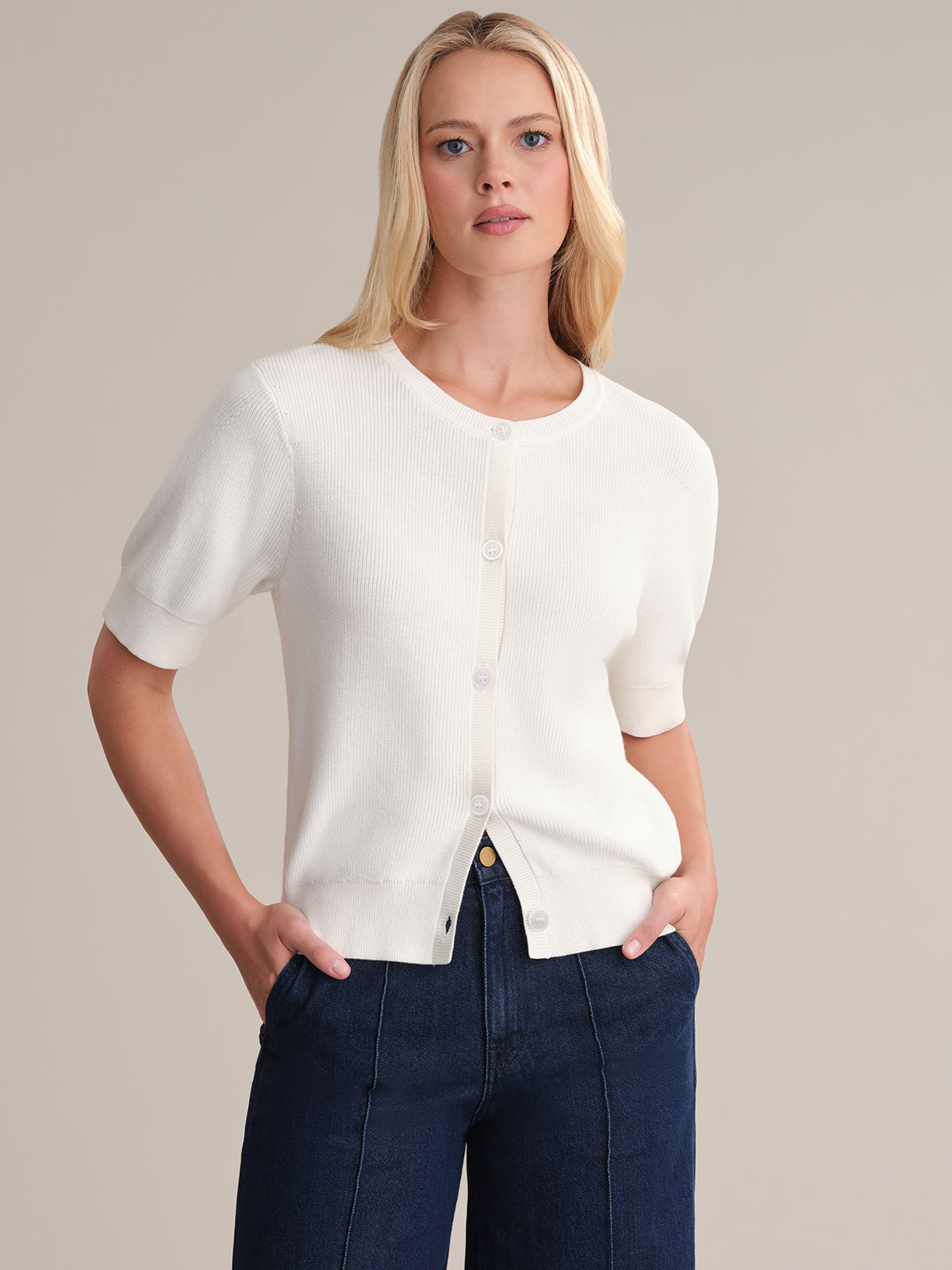 A person with long blonde hair is wearing a 525 America white short-sleeve puff cardigan, enhancing a relaxed silhouette. Paired with dark blue pants, they stand against a beige background, hands in pockets, showcasing the gentle drape of soft cotton fabric.