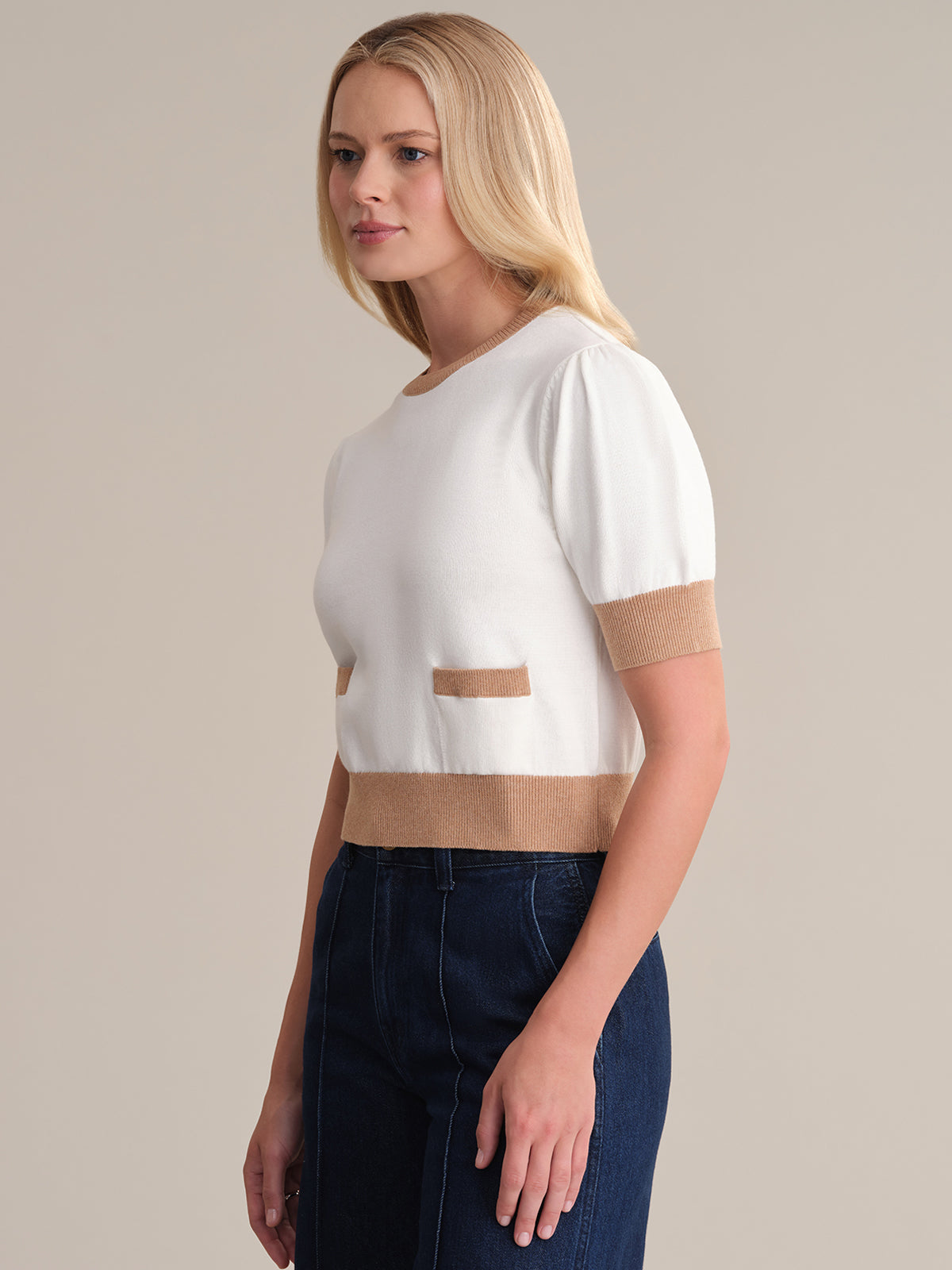 A blonde woman stands in profile against a neutral backdrop, wearing a 525 America pullover made of 100% cotton. It is white with brown-trimmed sleeves, pockets, and hem. She effortlessly pairs it with dark blue jeans.