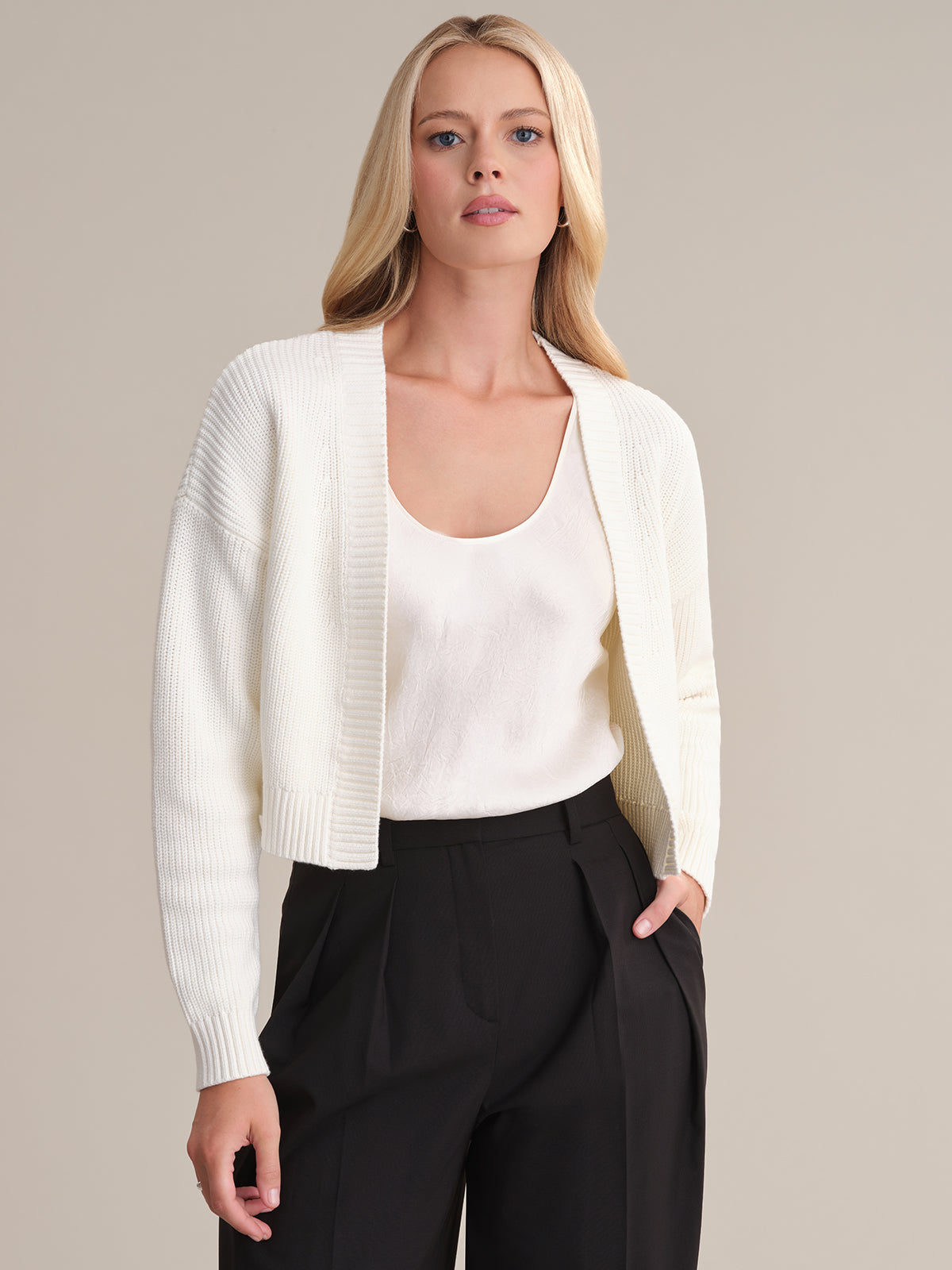 A person with long blonde hair wears a white, subtly ribbed cropped cardigan by 525 America over a white top and black pants. They stand against a plain beige background, one hand in their pocket, sporting a minimalist expression.