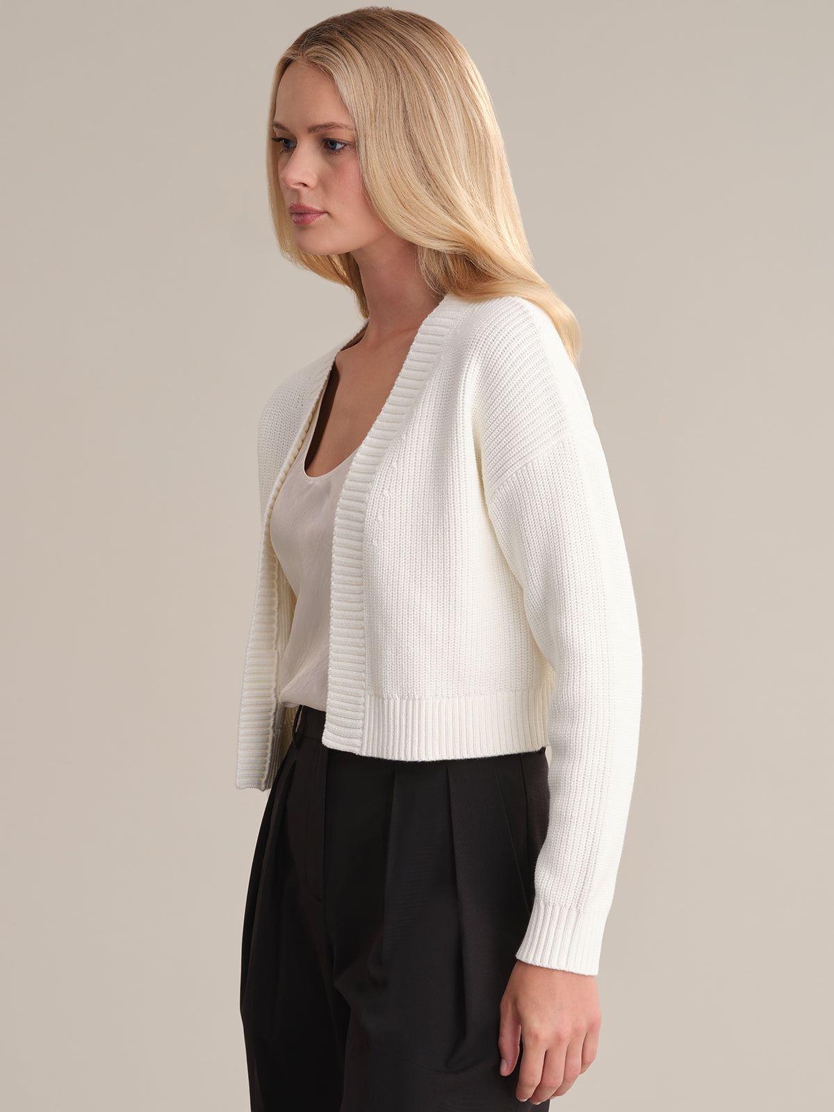 A person with long blonde hair is wearing a white, ribbed, cropped cardigan from 525 America over a white top and black pants. They stand against a minimalist background, looking to the side.