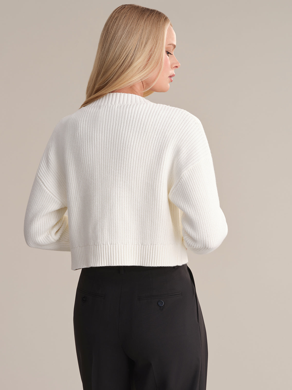 A person with long blonde hair is seen from the back, wearing a white ribbed sweater by 525 America and black pants. The minimalist design pops against the plain, soft beige background.