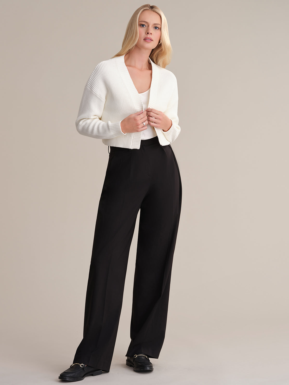 A person with long blond hair stands against a plain background, wearing a white ribbed cropped cardigan by 525 America over a white top, paired with black wide-leg trousers and black shoes. They maintain a neutral expression while looking at the camera, epitomizing minimalist design.