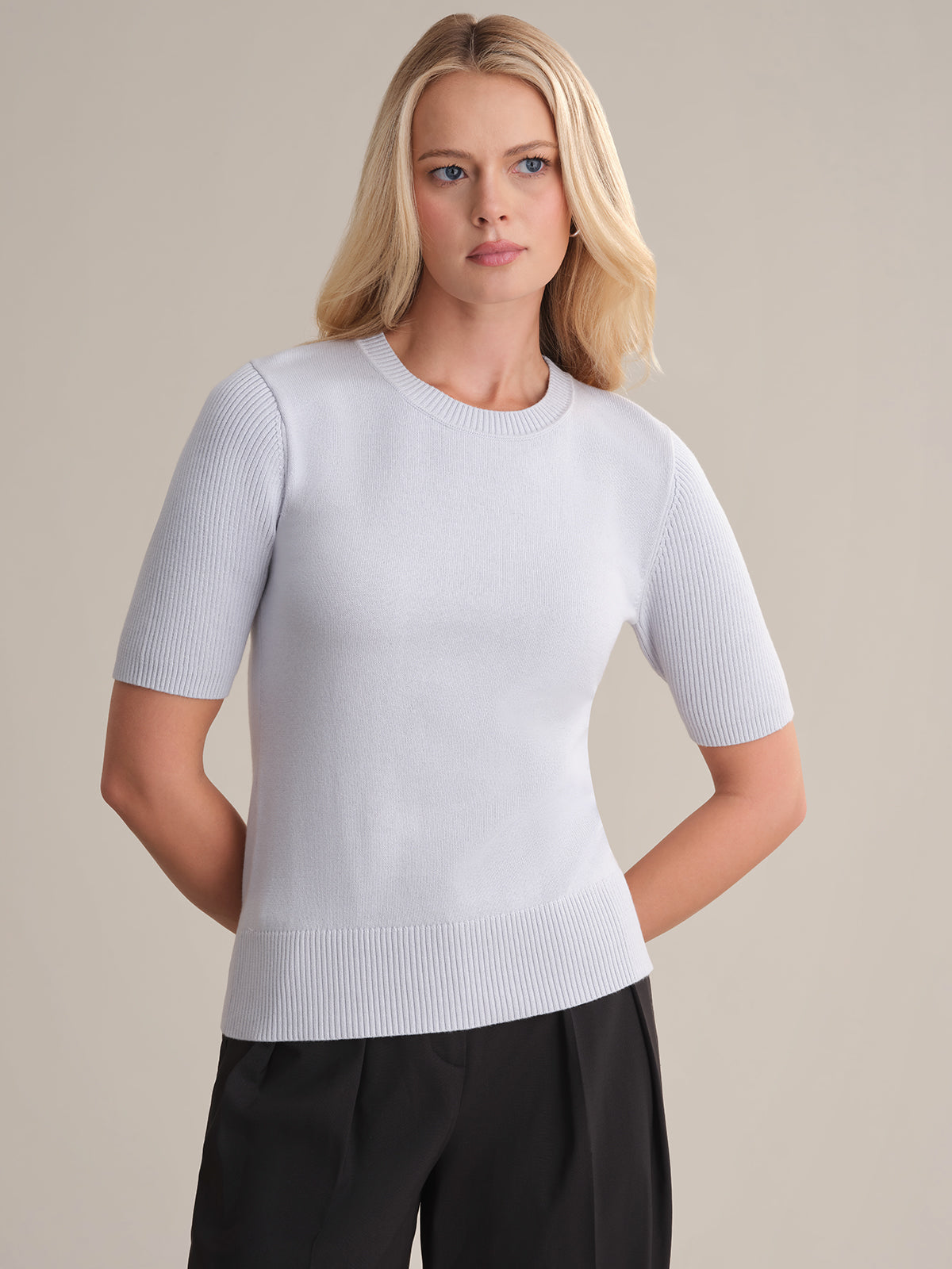A woman with long blonde hair wears a light gray 100% cotton Short Sleeve Cotton Sweater by 525 America with black pants, standing against a beige background—delivering effortless everyday style.