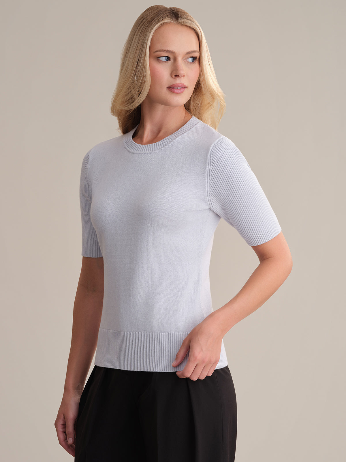 A person with light skin and long blonde hair wears a fitted, short-sleeve 100% cotton sweater by 525 America with black pants, standing against a beige background and looking to the side. Ideal for any everyday wardrobe.