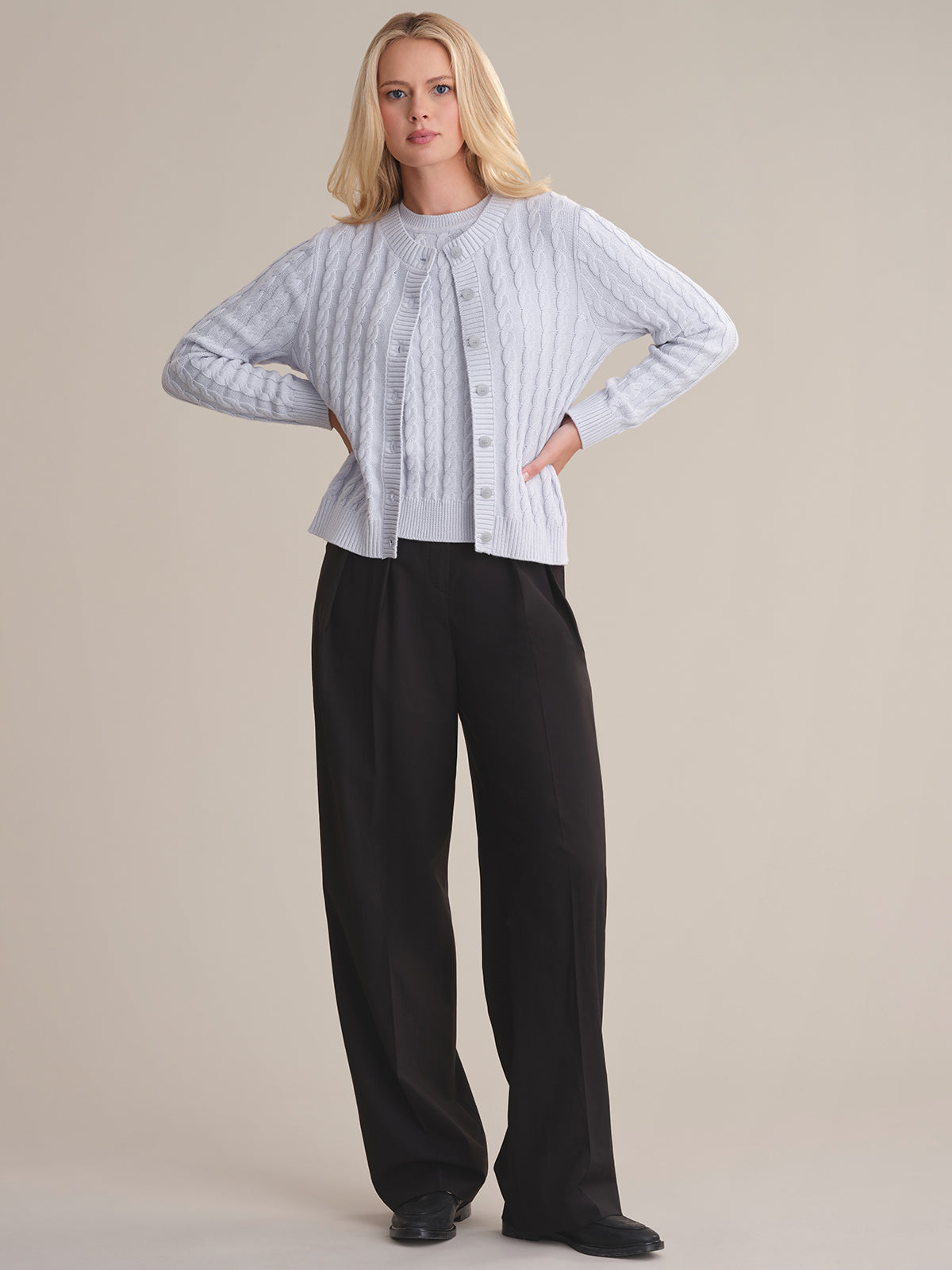 A person with long blonde hair and a neutral expression wears a light gray cable-knit cardigan by 525 America over a matching top, paired with loose black trousers and black shoes, standing against a plain background.