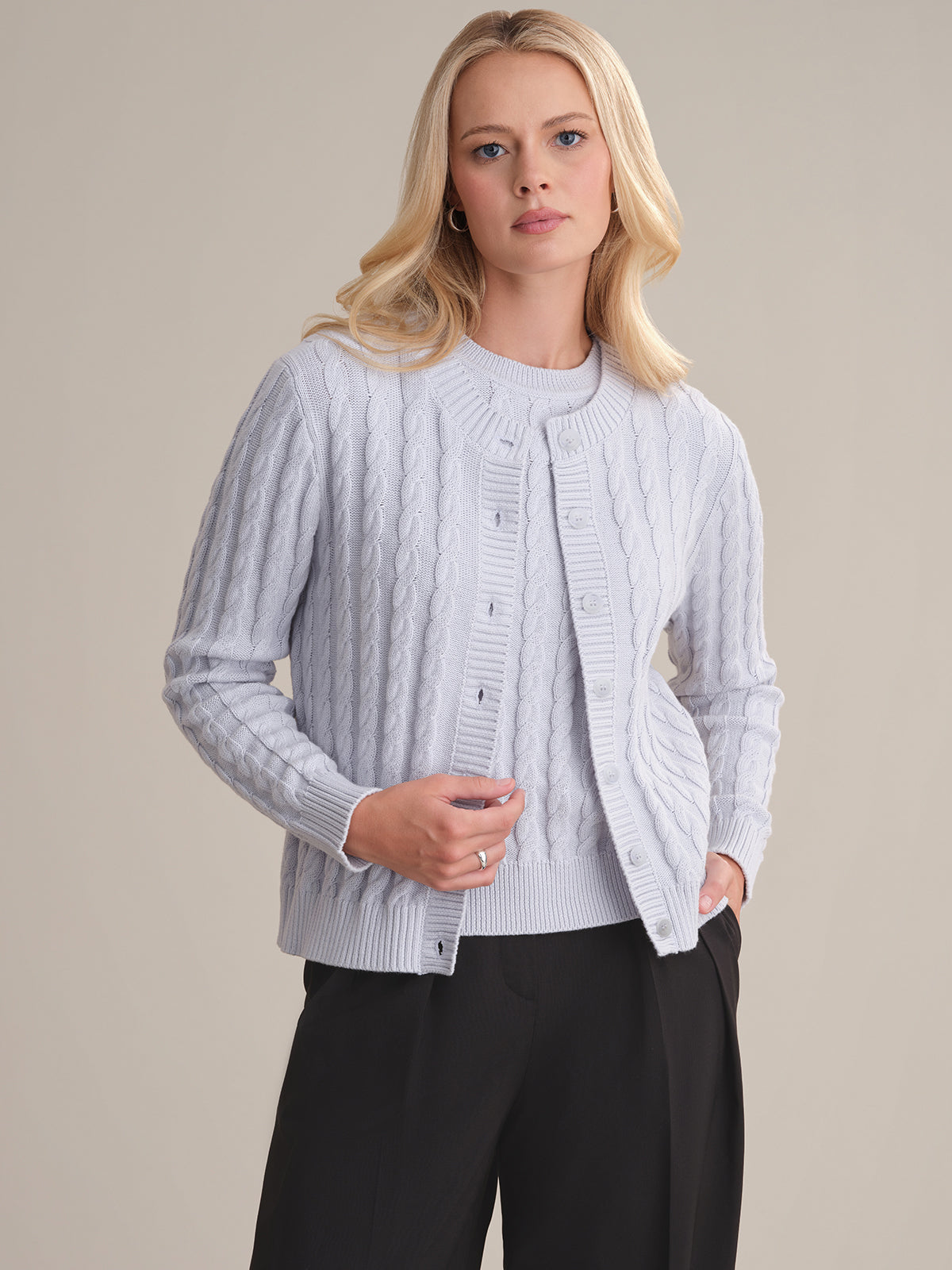A person with long blonde hair wears a 525 America light gray cable-knit cardigan and matching top with black trousers, standing against a plain light background, embodying the timeless appeal of classic fashion staples.