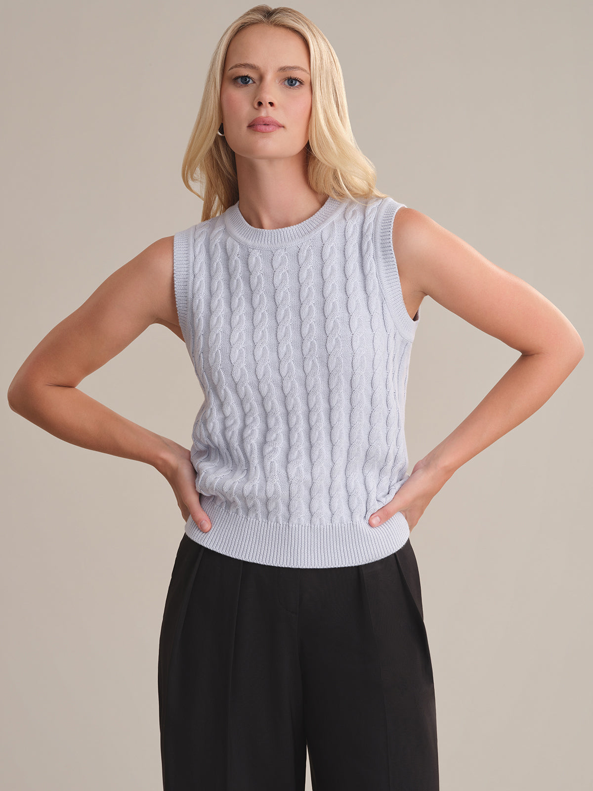 A person with long blonde hair models a 525 America light gray cable knit sleeveless sweater in a relaxed fit and black pants, standing against a beige background with hands on hips.