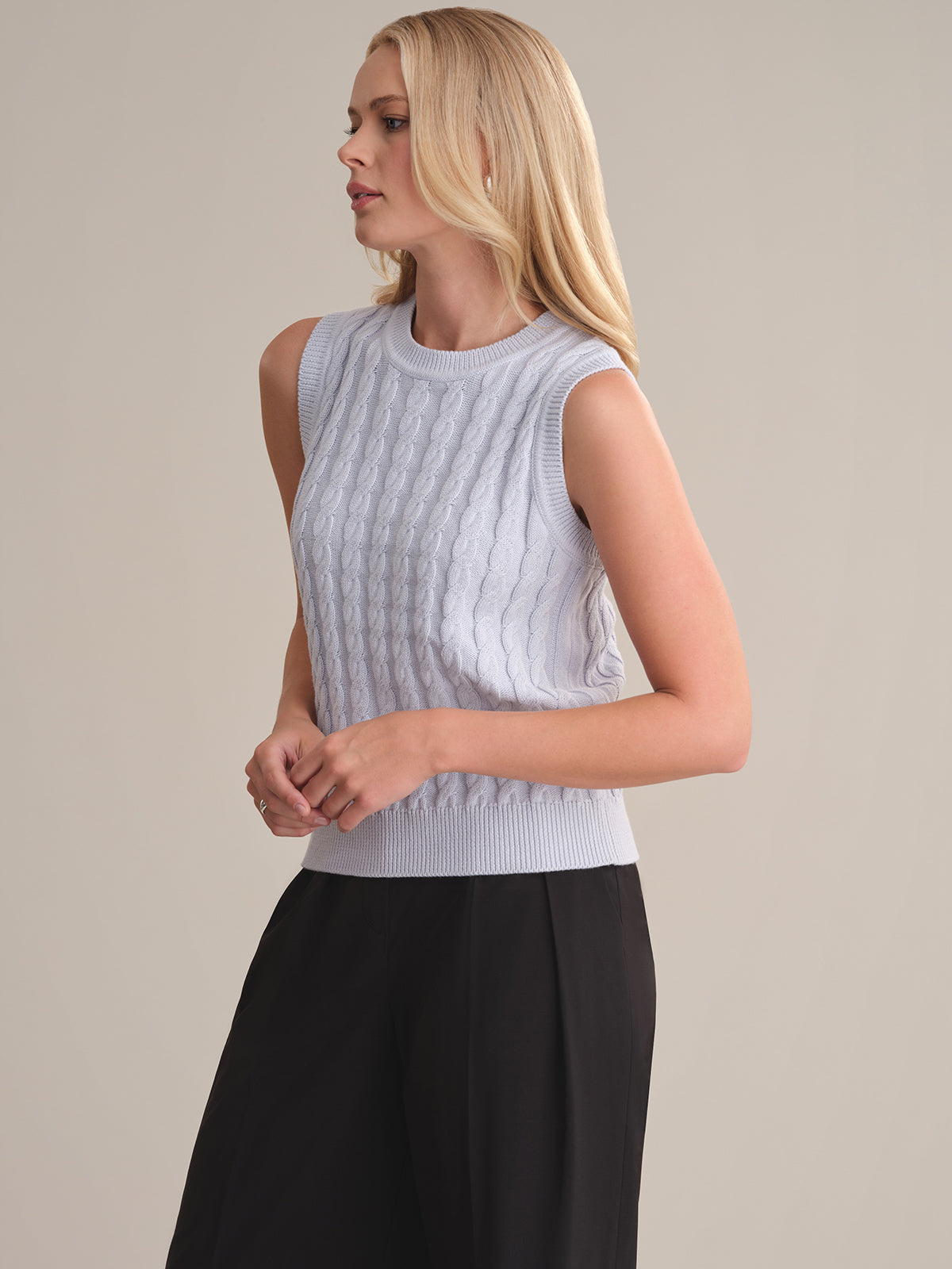A person with long blonde hair wears a 525 America light gray cable knit sleeveless sweater and black pants against a plain, light background, facing left with arms relaxed by their sides, highlighting the sweaters relaxed silhouette.