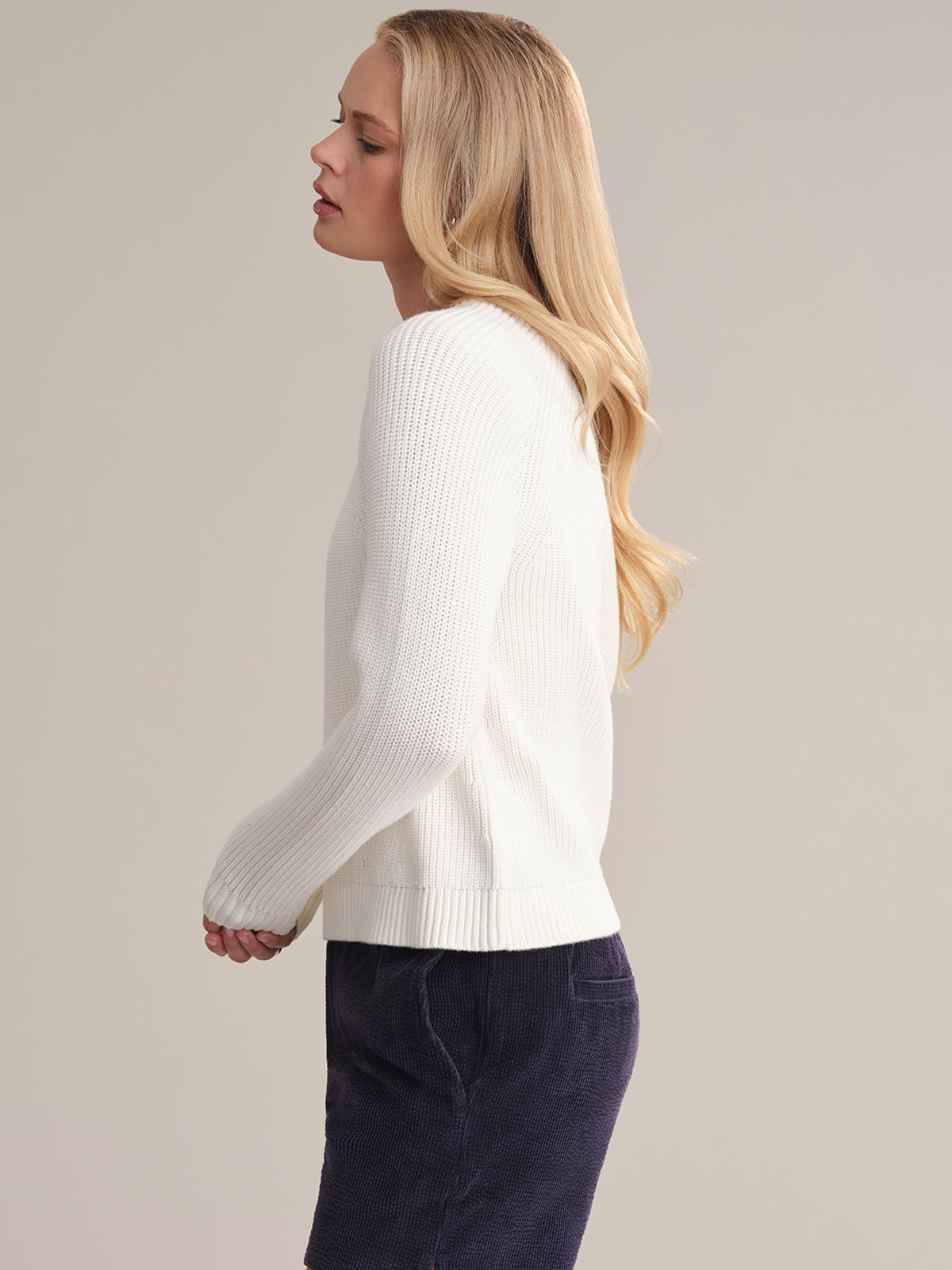 A person with long blonde hair is shown in profile against a beige background, wearing a fitted white cotton shaker stitch sweater with raglan sleeves by 525 America, paired with dark pants.