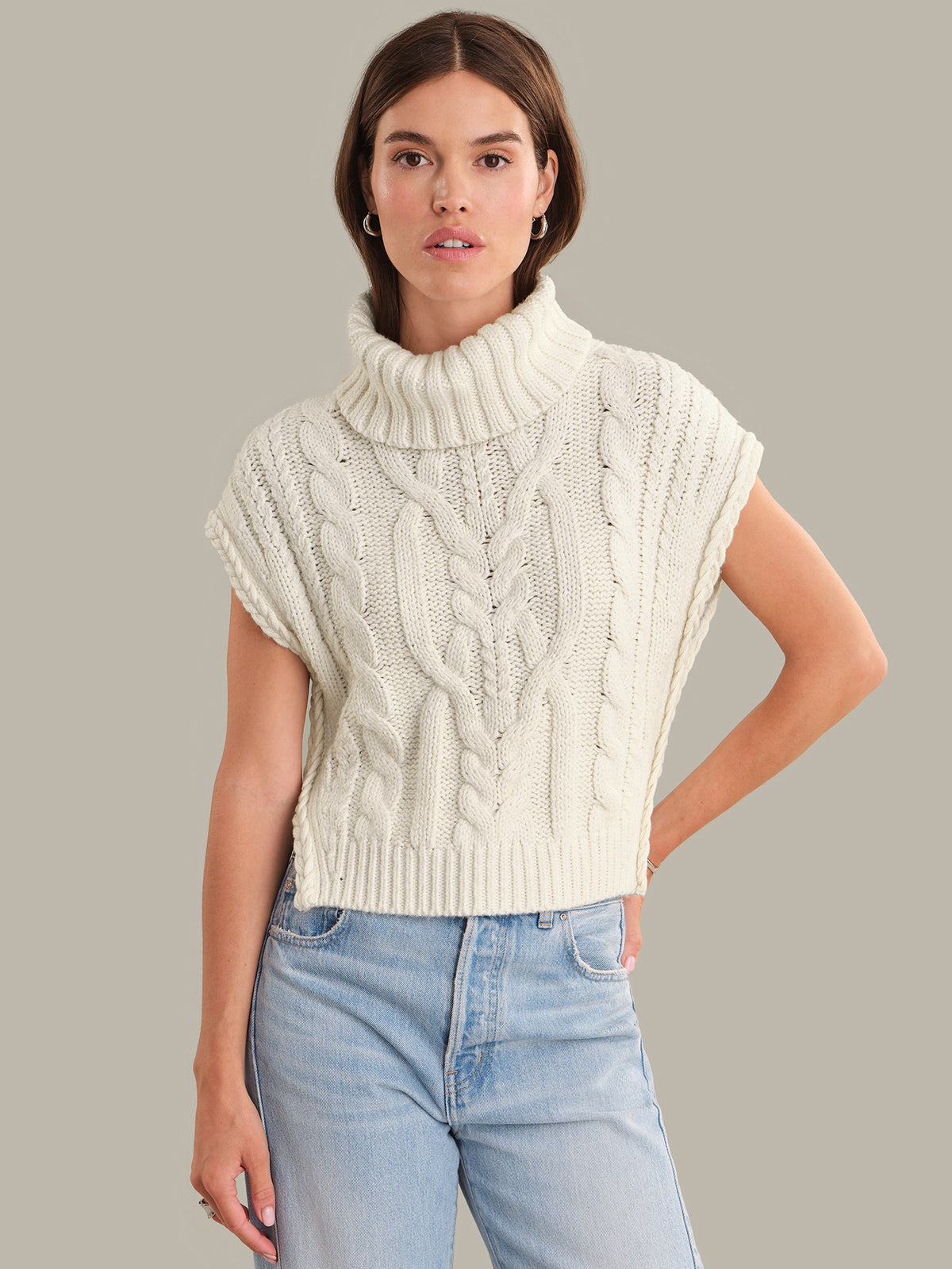 Woman wearing a cream-colored knitted sleeveless sweater with light blue jeans on a gray background