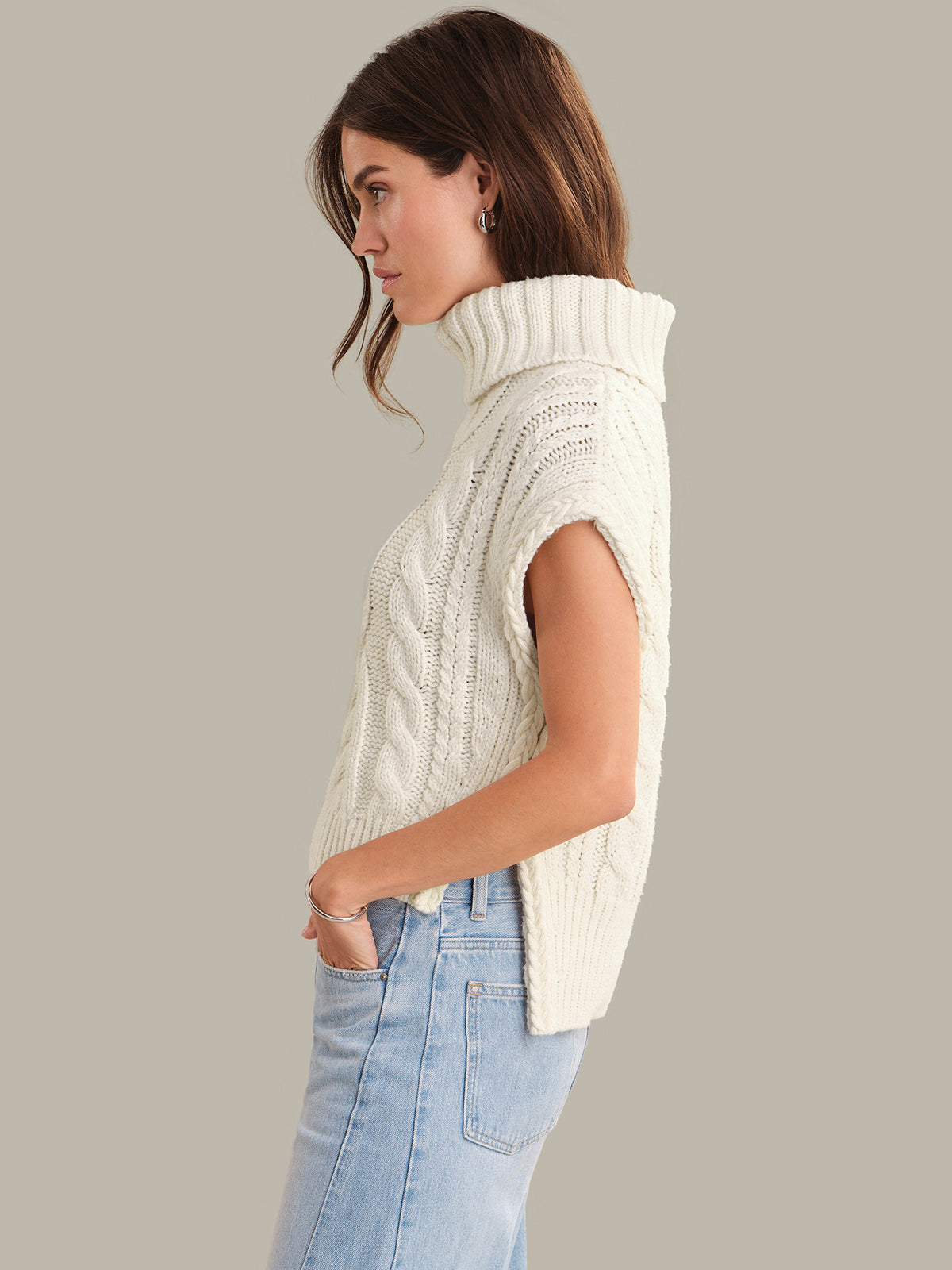 Woman wearing a cream sleeveless knitted sweater against a plain background
